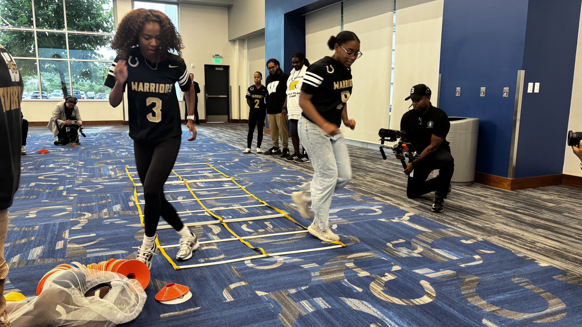 Indoor training: athletes in black "Warriors" jerseys on a blue gym floor. Foreground player (number 3) runs a ladder drill as a cameraman films from the right and others watch by blue walls.