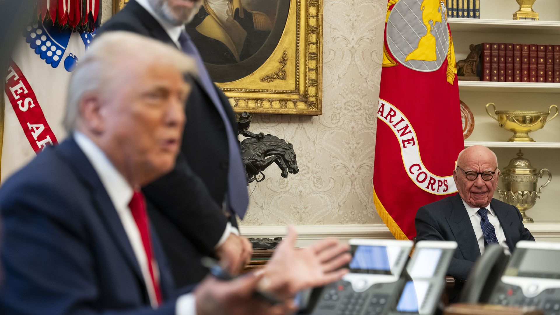 An older man in glasses and a suit sits near a red Marine Corps flag and trophies, while a blurred person in a suit gestures in the foreground in an official-looking room with books and a painting.