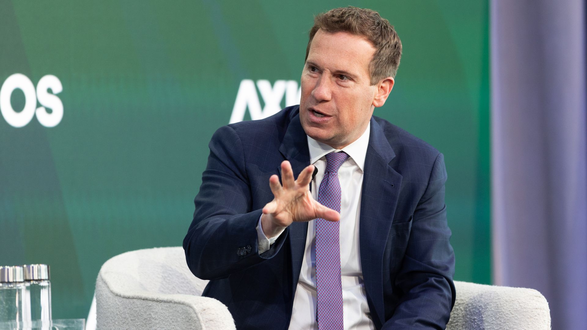 A man in a navy suit and purple tie sits in a light gray armchair on a stage, gesturing with his left hand as he speaks; green backdrop with partial white letters in the background.