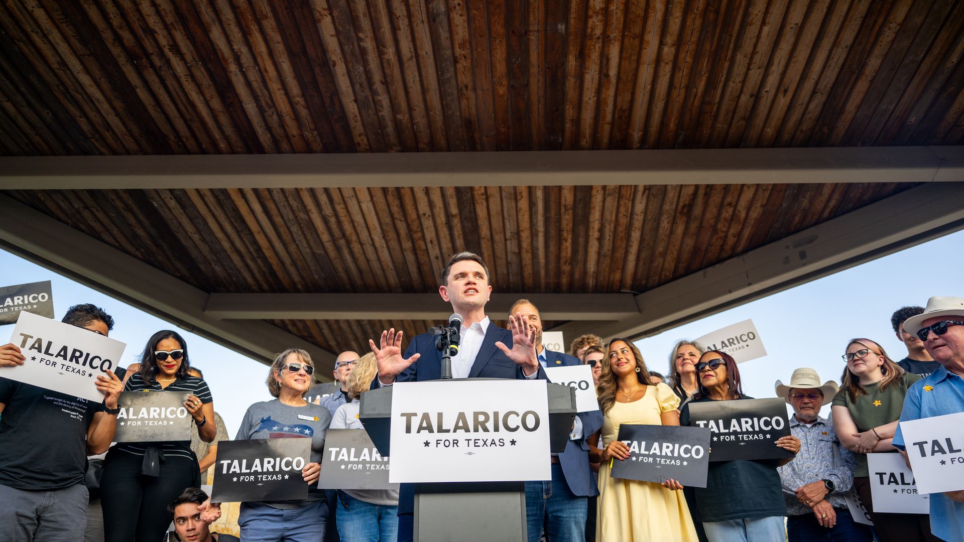 James Talarico stands behind a podium with a "Talarico" campaign sign, speaking with several supporters behind him.