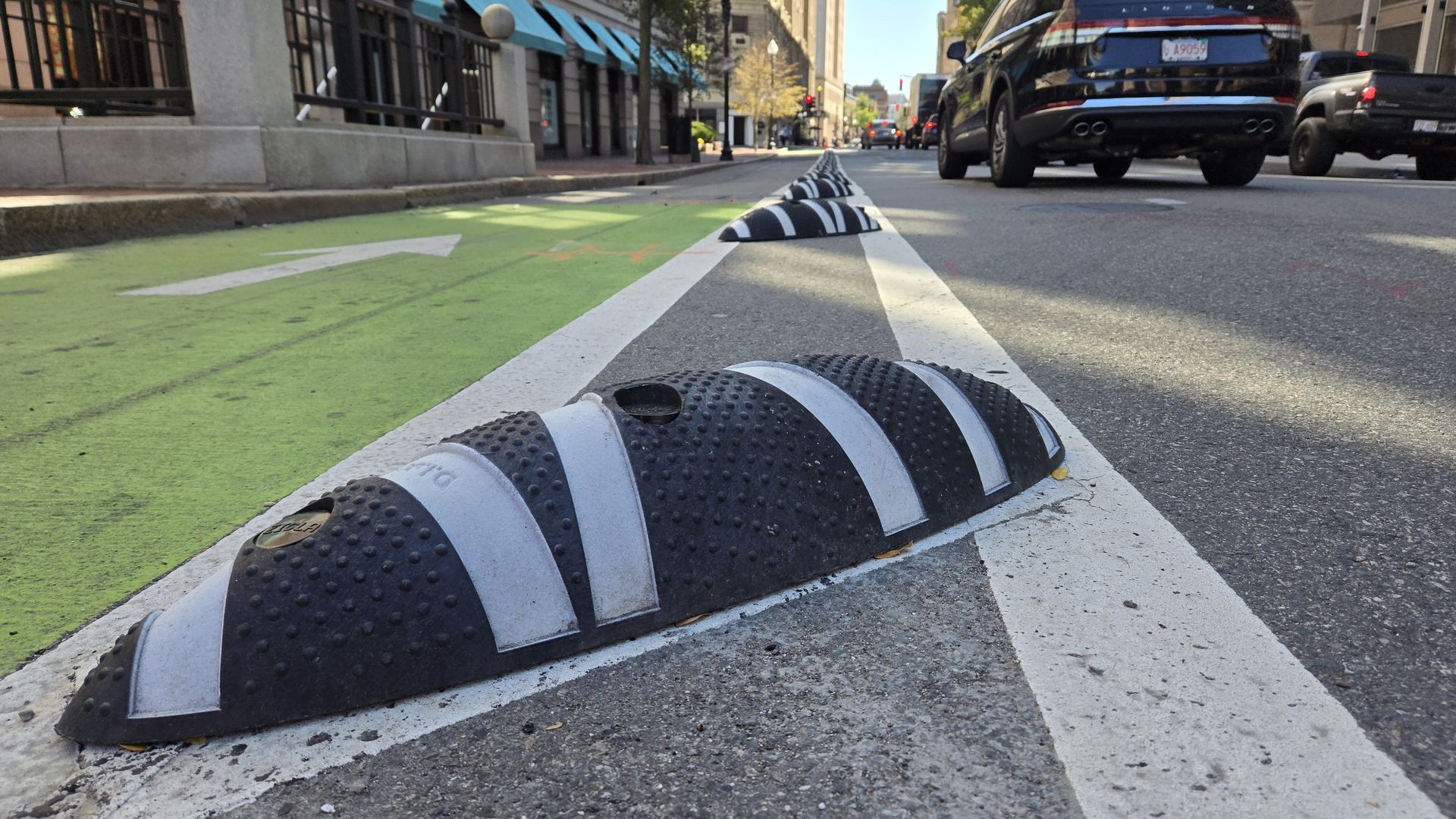 Close-up of black and white striped road separators on a city street dividing a green bike lane and a vehicle lane with cars and buildings in the background