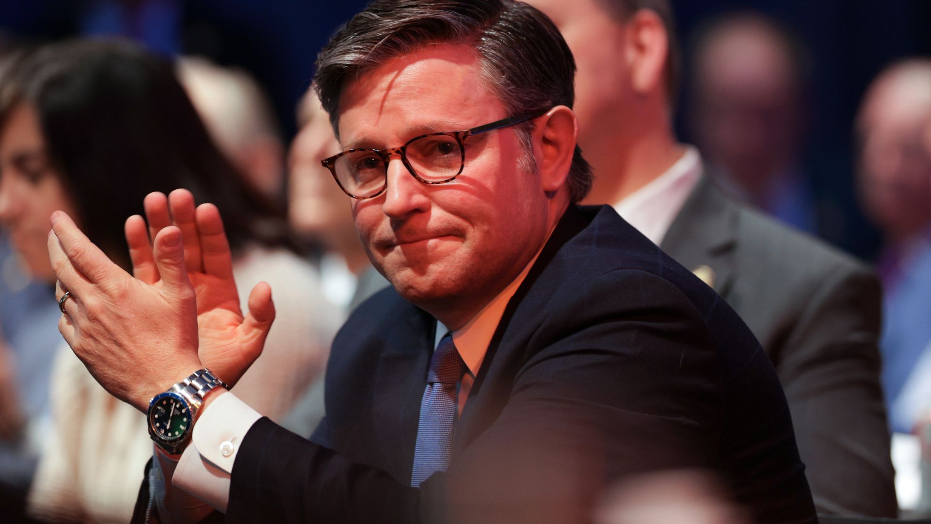 Man with glasses in a dark suit and blue tie clapping, seated among a group of people, with soft lighting and blurred background.