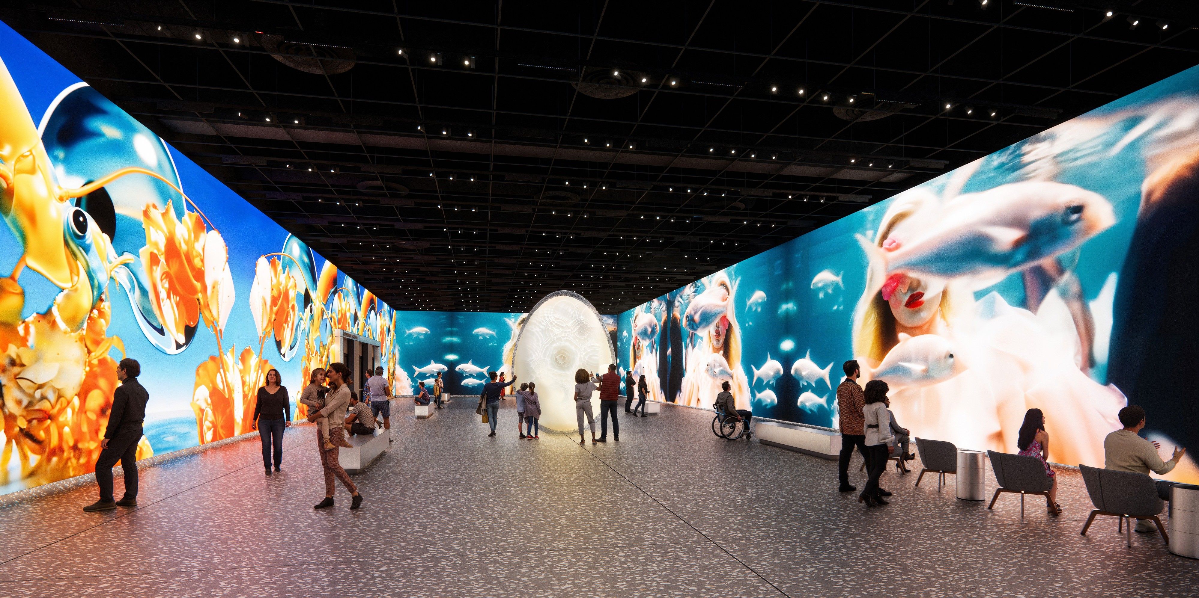 People exploring a digital art gallery with large vibrant screens showing orange-yellow abstract sea creatures on one side and underwater scenes with fish and a woman on the other.