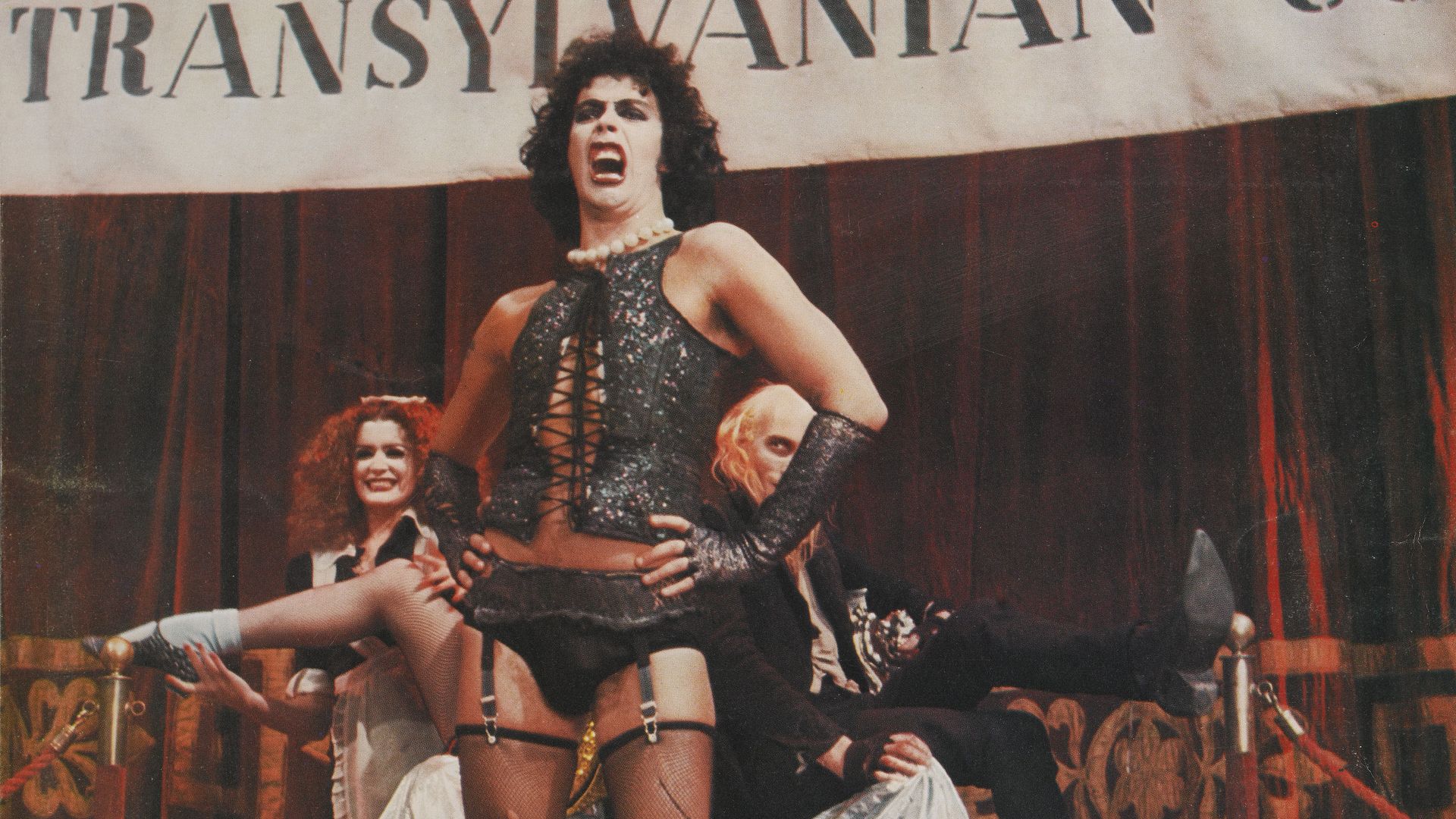 Tim Curry in a bustier and black underwear with garters, with hands on hips, as Frank-N-Furter in a still from The Rocky Horror Picture Show.