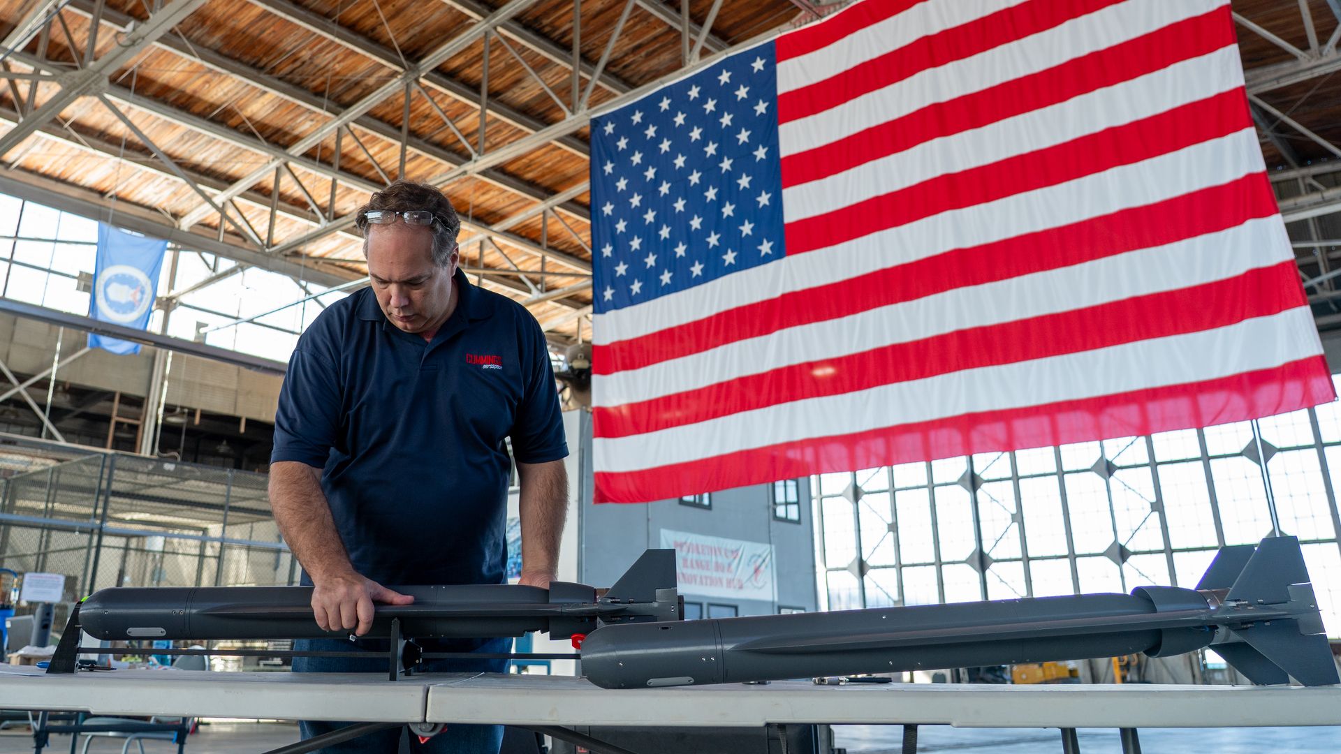 A man in a dark blue polo shirt works on a gray rocket-like device on a table inside a large hangar, with a big American flag hanging in the background.