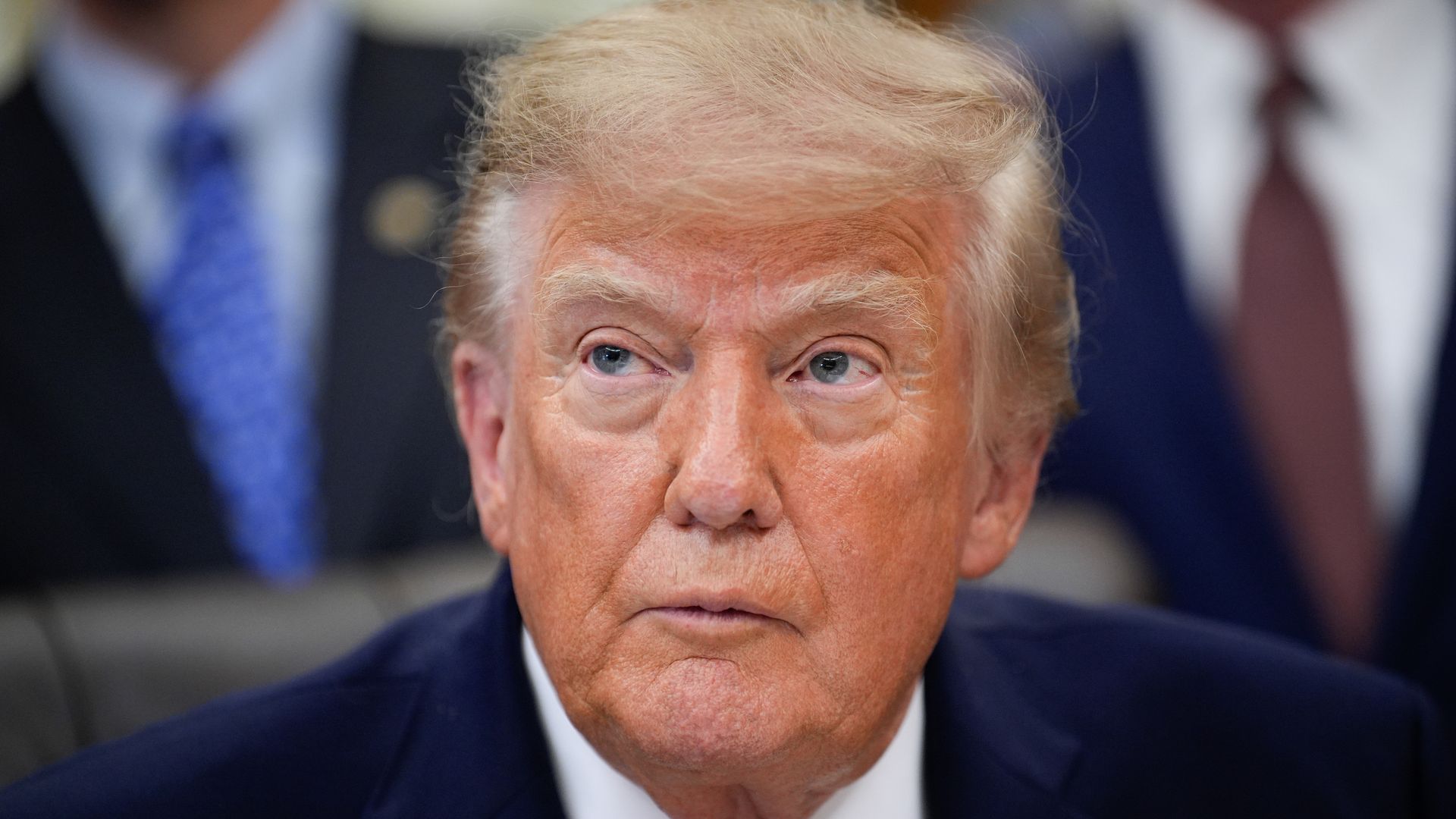 An up-close shot of President Trump sitting in the Oval Office while wearing a white collared shirt, a dark blue jacket, a red tie and an American flag pin.