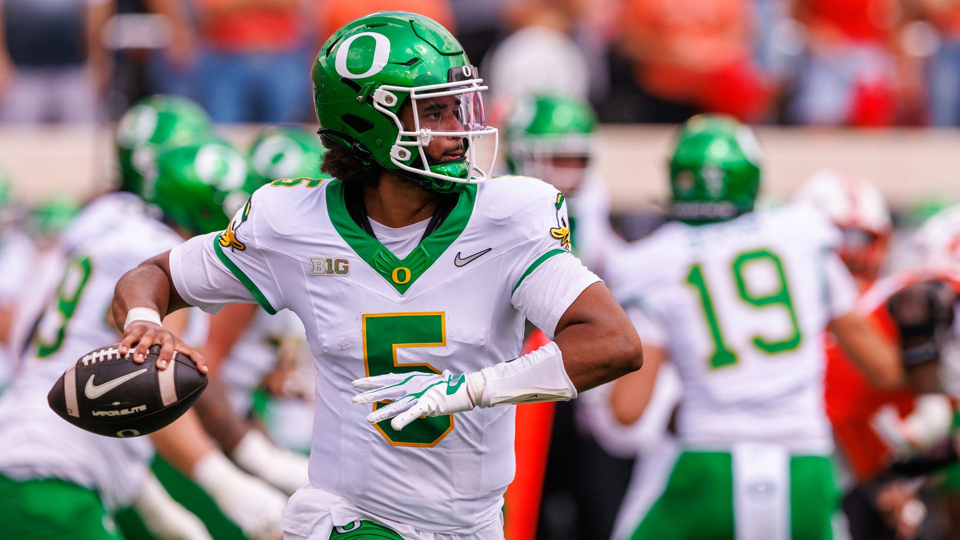 A college football player in a green Oregon helmet with a white O runs forward, holding a football, while teammates in white and green surround him and a blurred crowd behind.