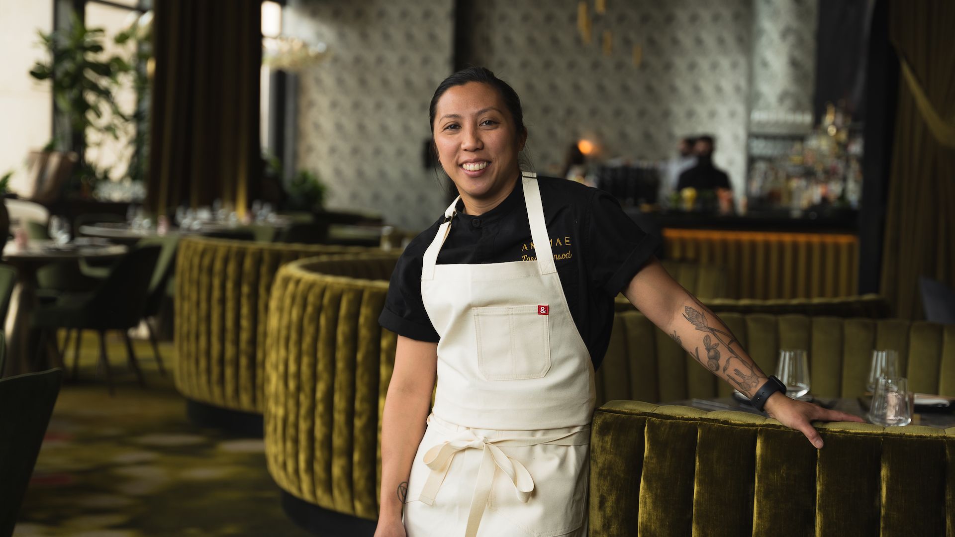 Chef Tara Monsod wears a white apron and leans on a booth in the restaurant Animae.