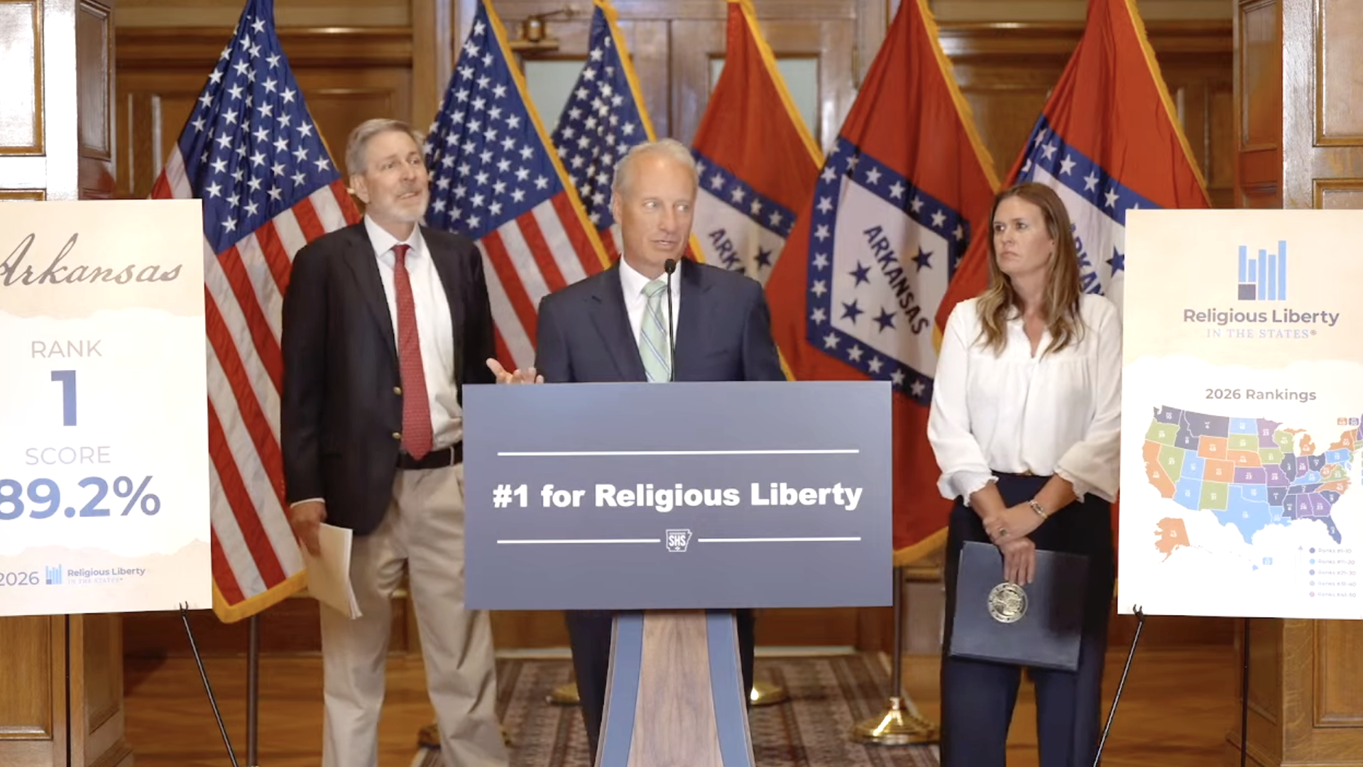 A speaker stands at a podium during a news conference announcing Arkansas' top ranking for religious liberty, flanked by officials, U.S. and Arkansas flags, and ranking display boards.