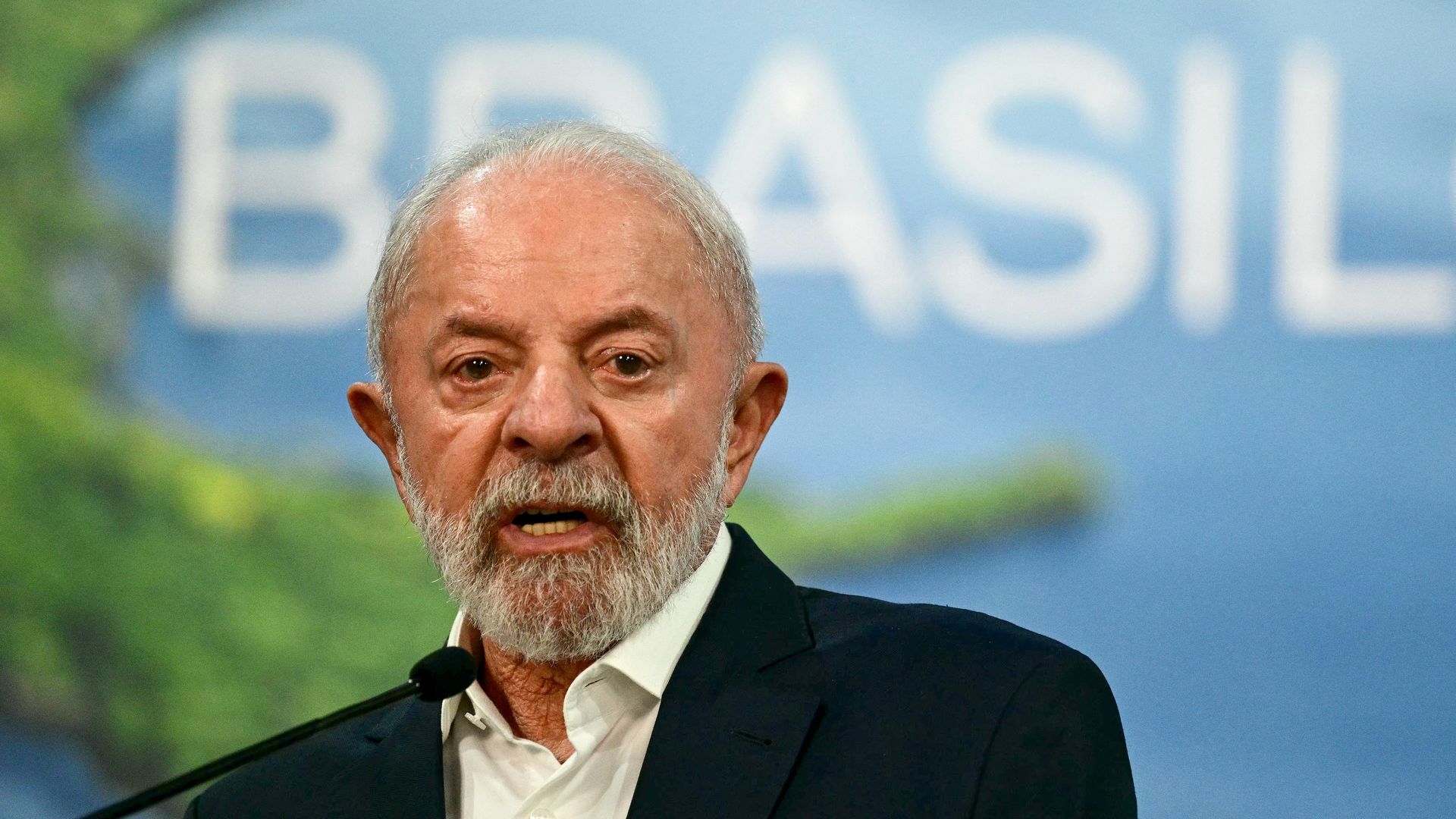 Close-up of Brazil's President Luiz Inacio Lula da Silva, a bearded older man in a dark suit speaking at a podium, wearing a lapel microphone. A blue backdrop with large white letters is behind him.