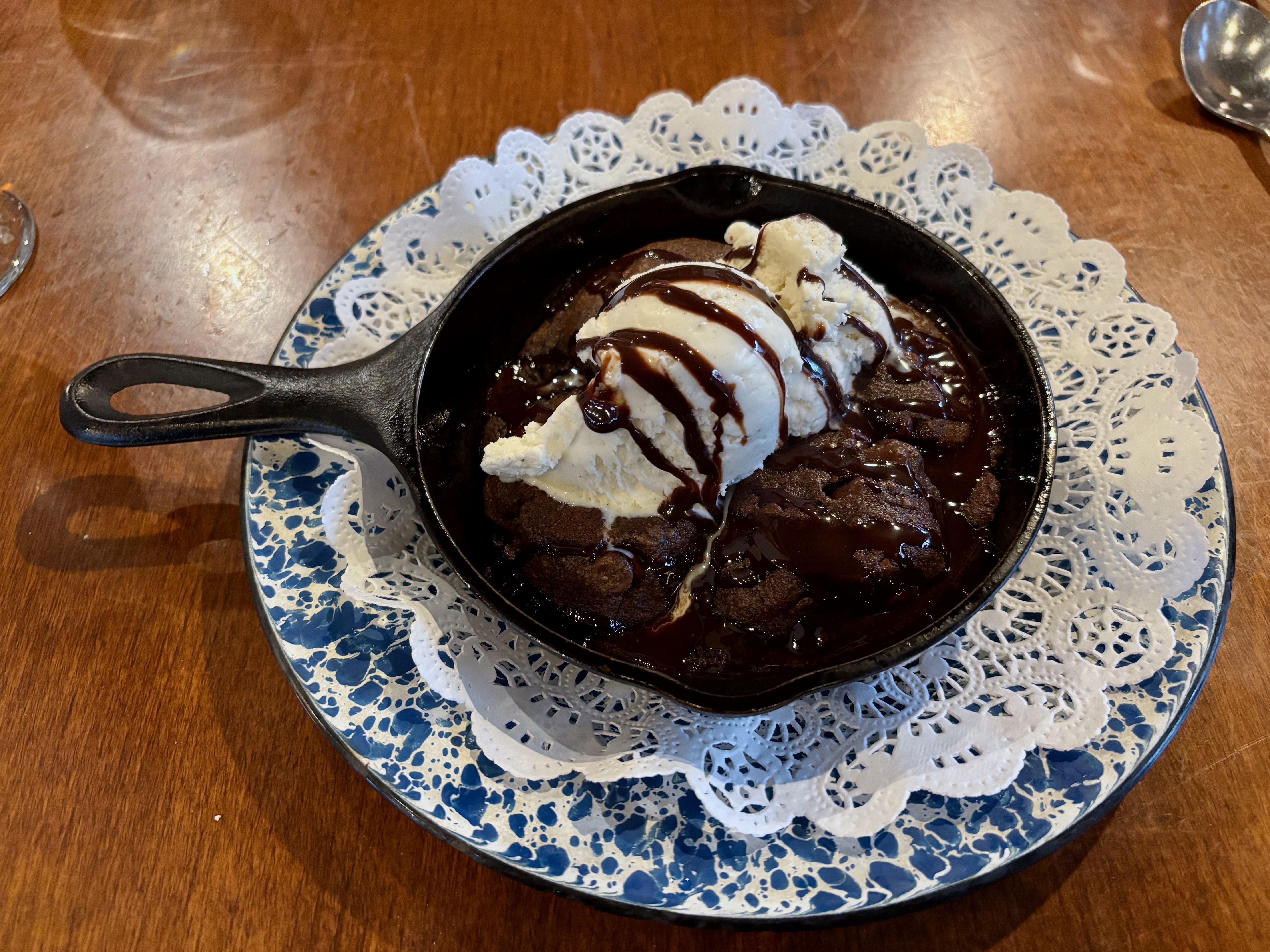 Skillet chocolate brownie topped with vanilla ice cream and chocolate sauce, served on a blue and white plate with a lace doily under the skillet, on a wooden table.