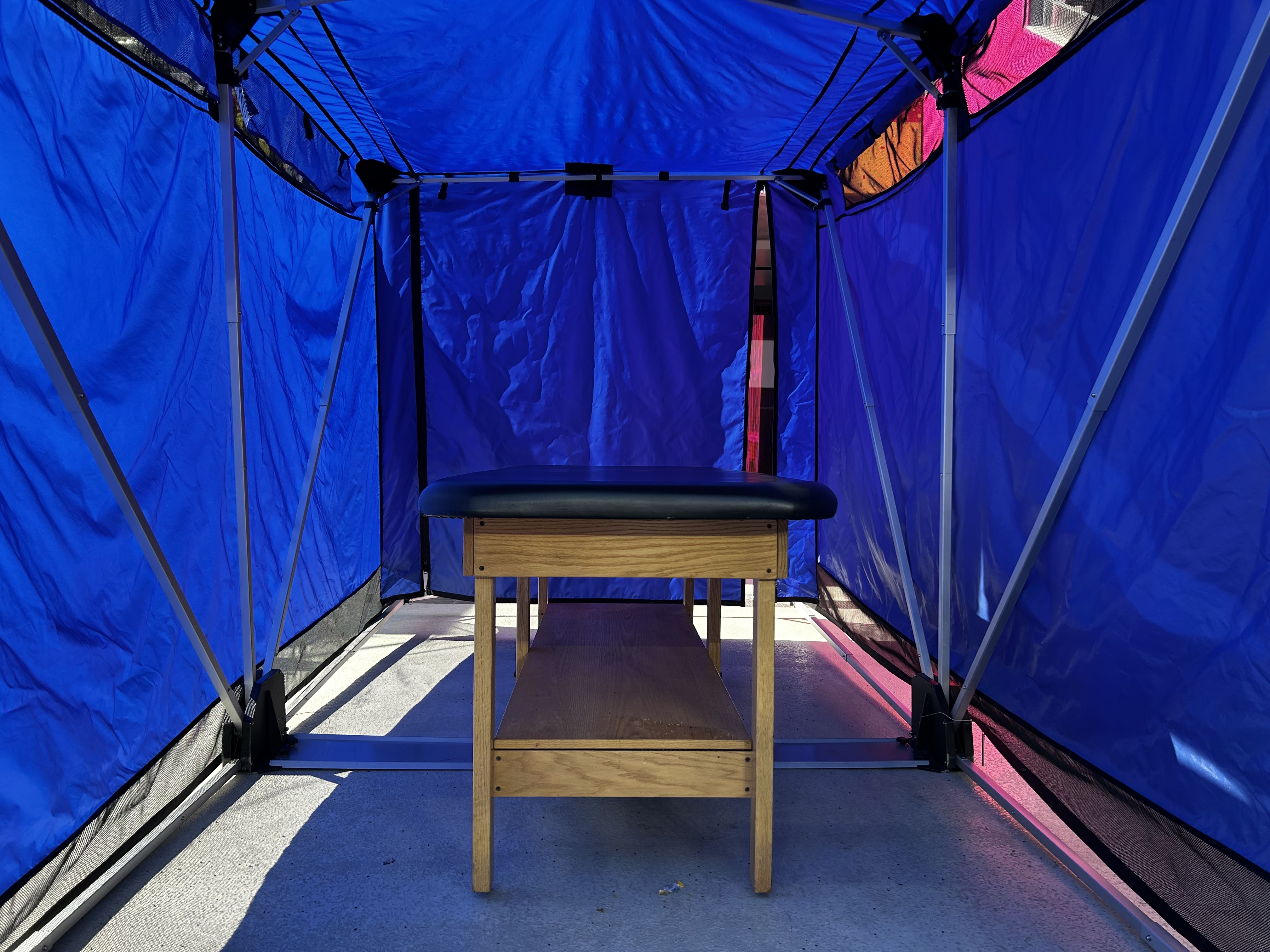 Wooden massage table with black cushioning inside a blue fabric-covered canopy tent on a concrete floor with sunlight and shadows.