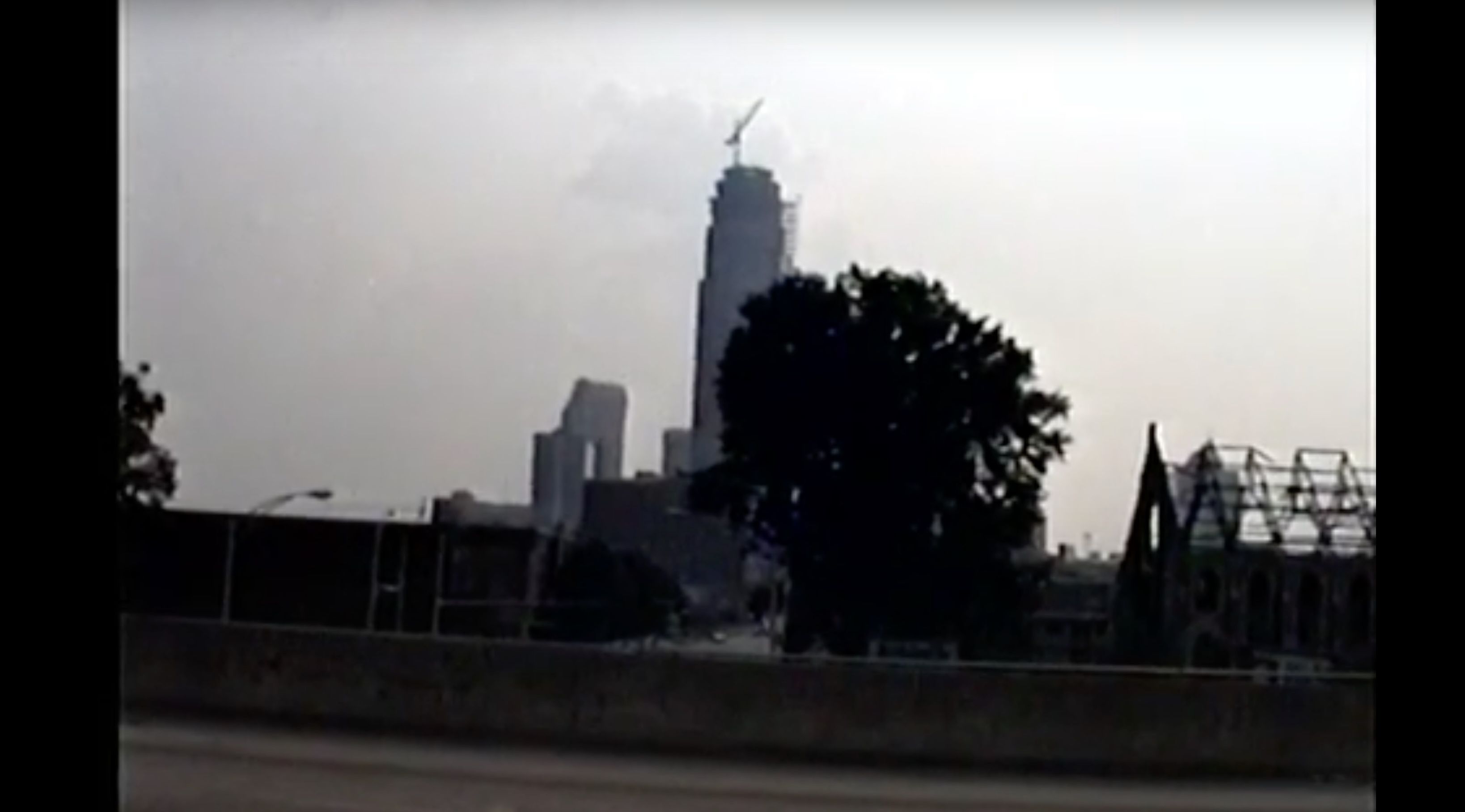 Cityscape with a tall building under construction and a crane on top, a large tree silhouette in the foreground, and an incomplete metal roof structure on the right under a gray sky.