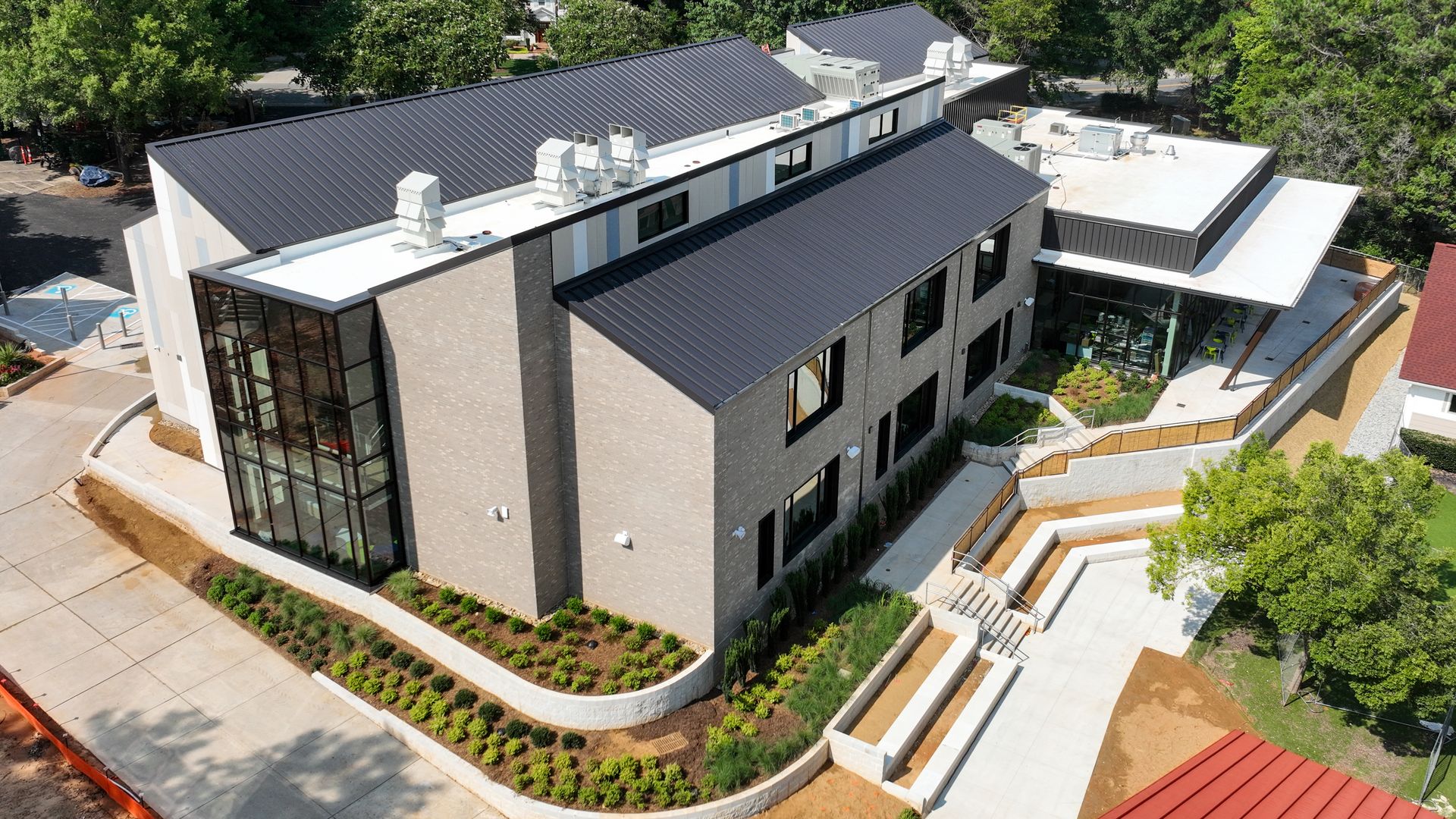 A modern two-story building with gray brick walls and dark metal roofs, large glass windows, surrounded by landscaped greenery and paved walkways, set amid tall trees and nearby houses.