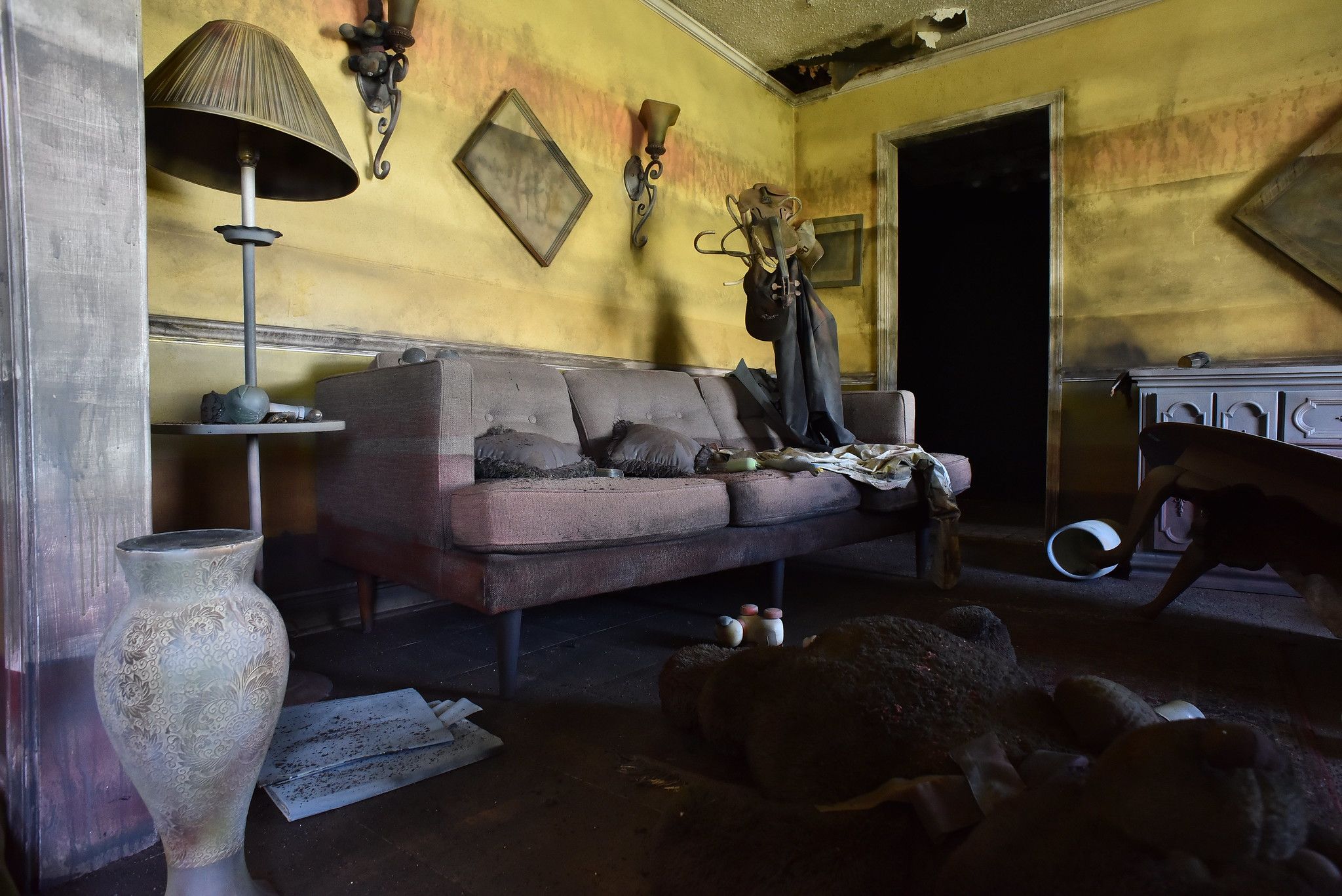 Dimly lit, dusty vintage living room with yellow-stained walls, worn brown sofa, old lamp, a broken teddy bear on the floor, and a torn ceiling with peeling paint.