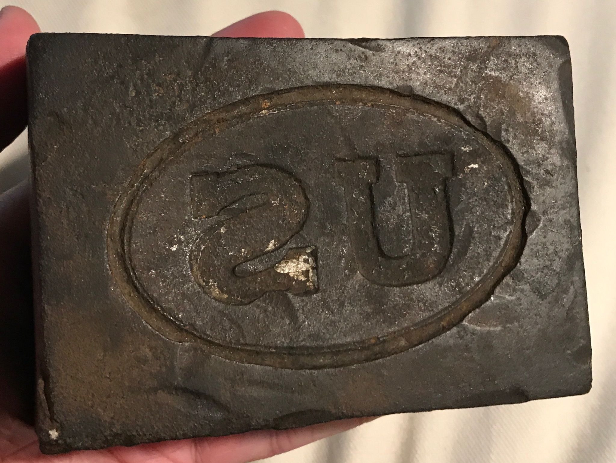 Close-up of a dark, weathered stone seal with a carved circular emblem. An oval ring contains abstract curved lines; the surface shows dust and white specks.