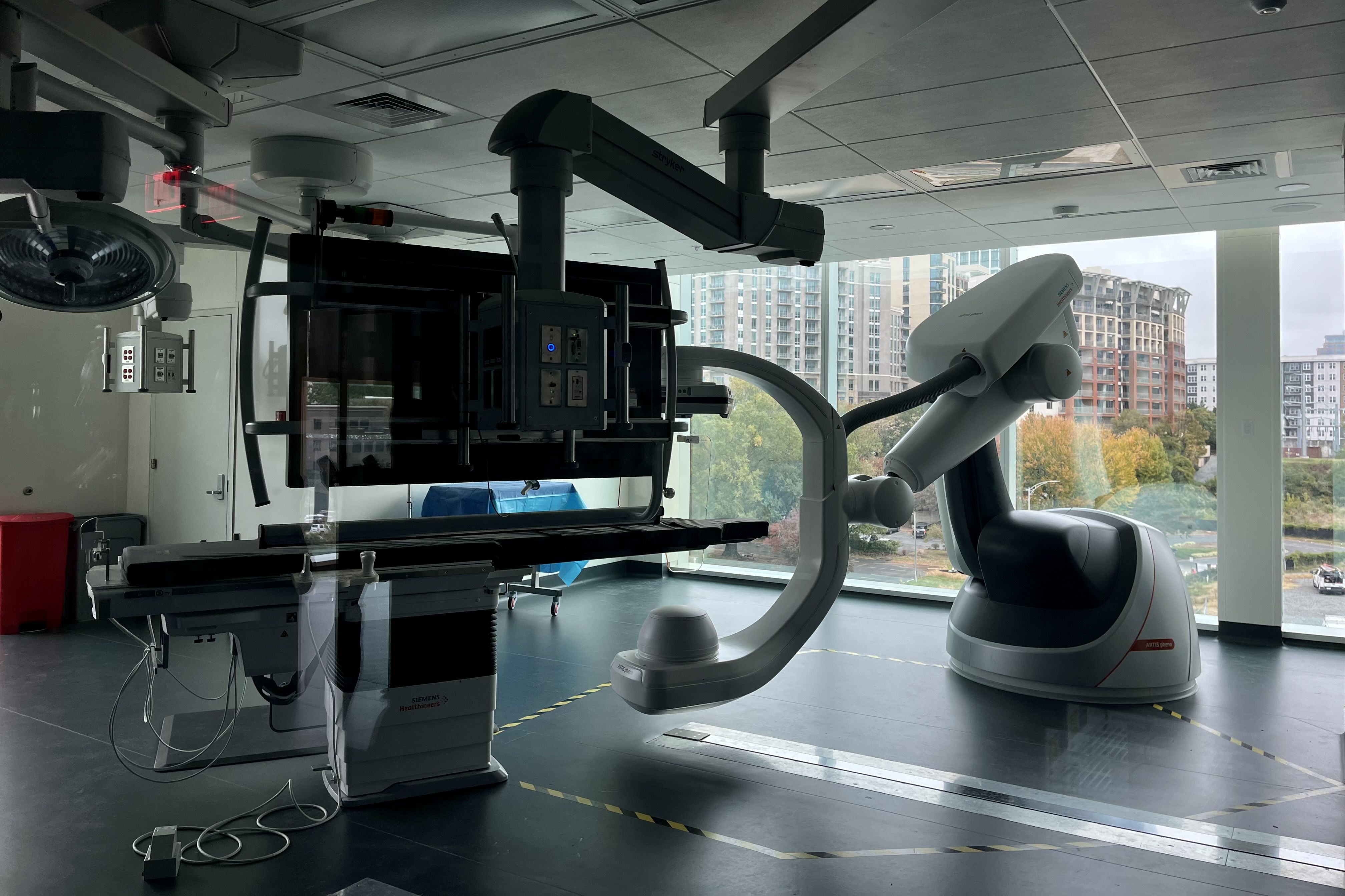 Modern medical imaging room with Siemens Healthineers equipment, featuring a robotic arm and adjustable patient table, large windows show city buildings and green trees outside.
