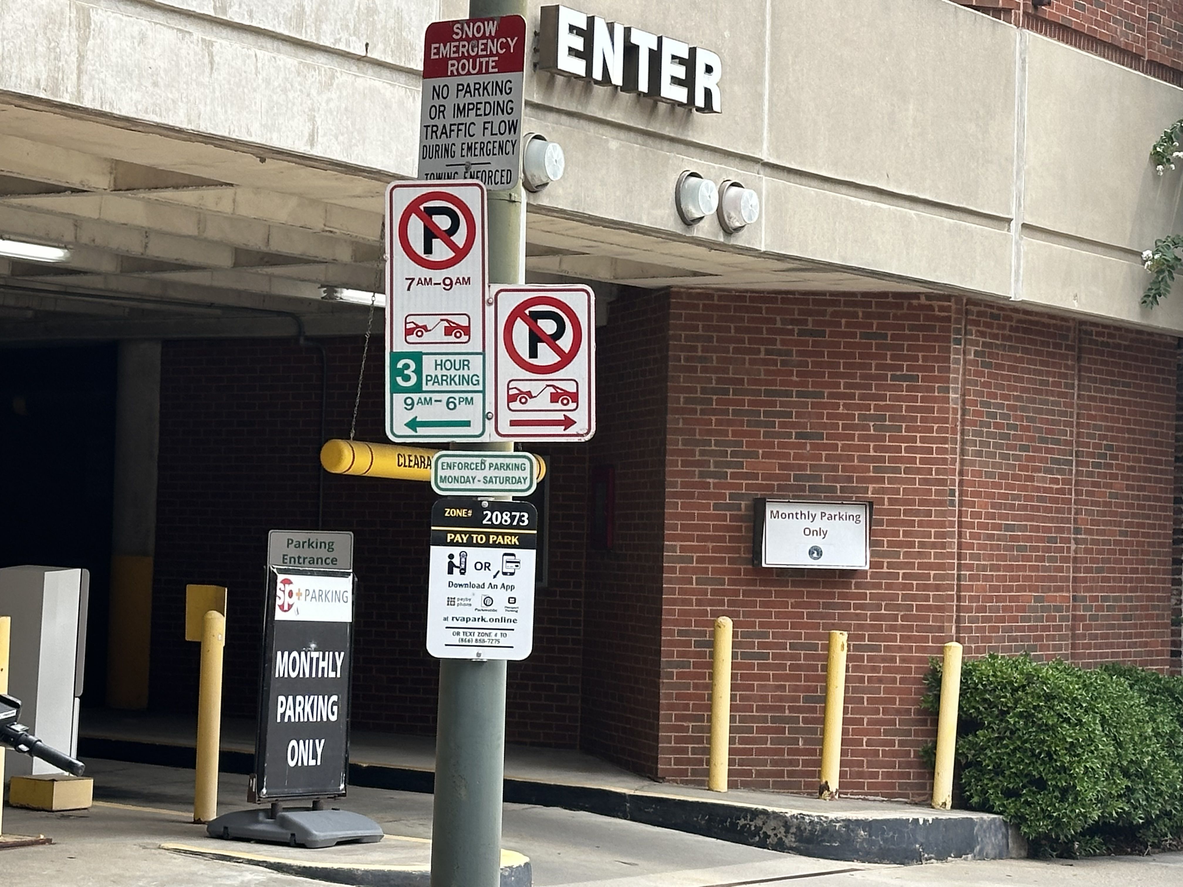 Parking garage entrance with signs reading "Snow Emergency Route" and "No Parking", plus "Hour Parking 9 AM-6 PM". Large "ENTER" on the facade, with yellow bollards and brick walls.