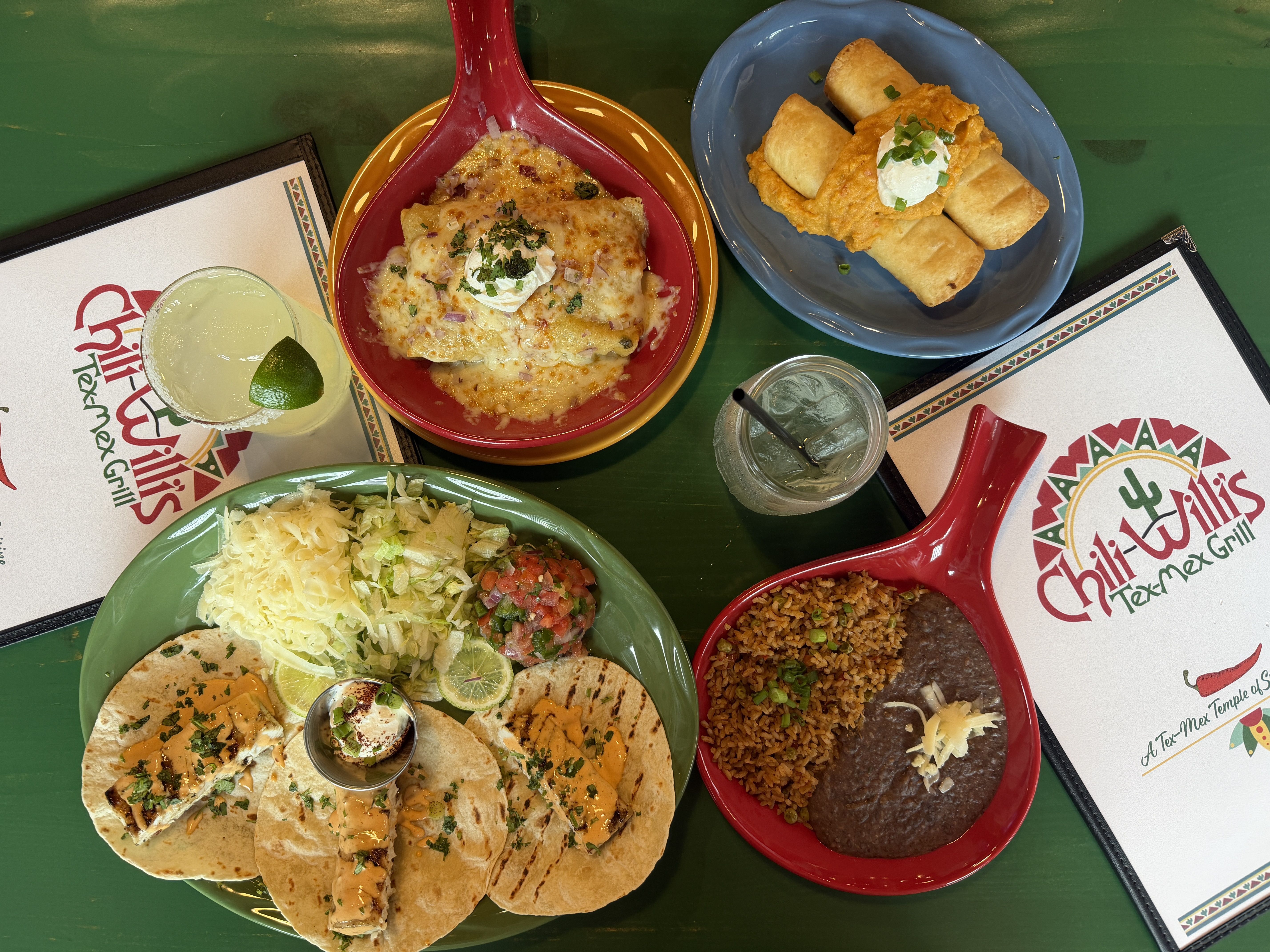 Top-down view of a Tex-Mex feast: red skillet of cheesy enchiladas with sour cream, blue plate with taquitos and sour cream, green rice, and tacos with sauces on a green table.