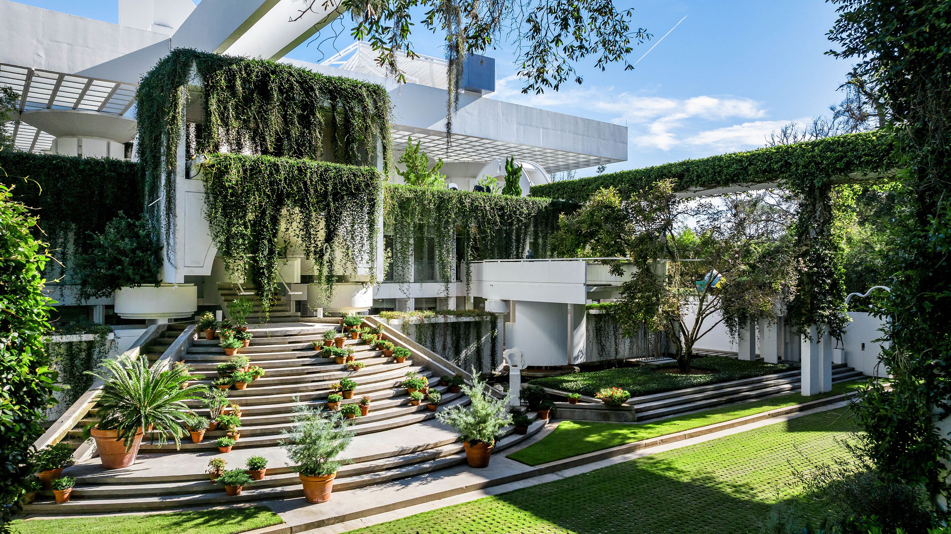 Vines hang from the balconies and roof of a large neo-futurist white beachfront mansion 
