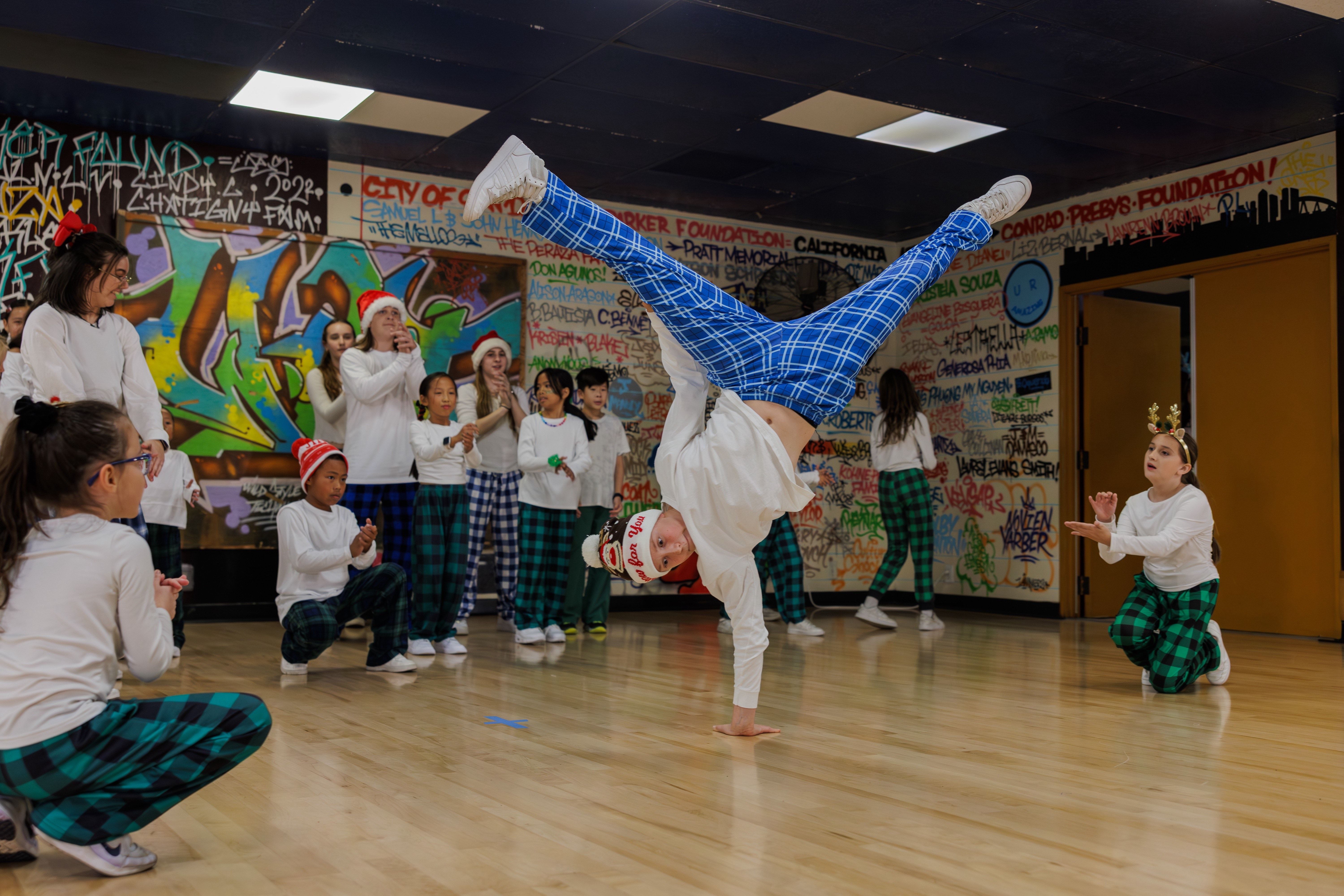 Child in blue plaid pants and white shirt doing a handstand in a room with graffiti walls, watched by a group of kids in holiday hats and white shirts, some clapping and cheering.