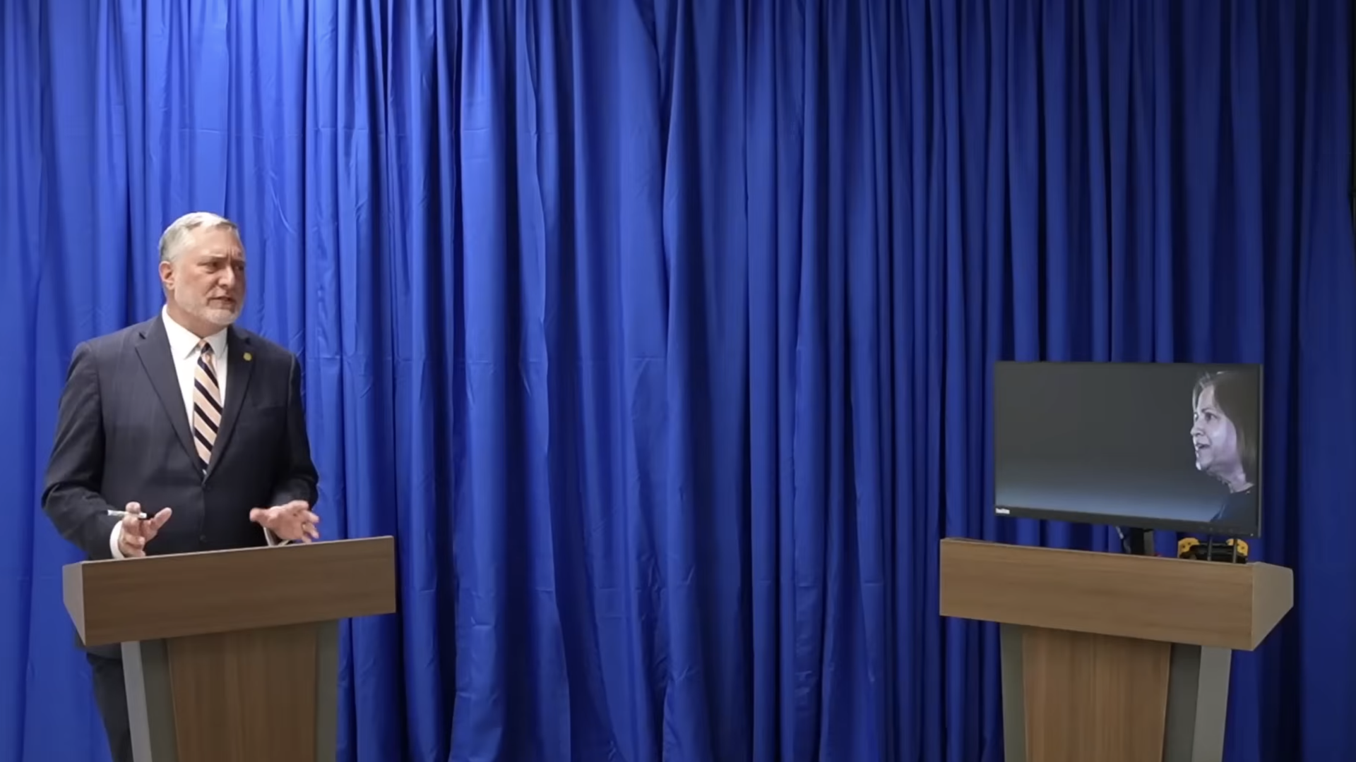 Man in dark suit and striped tie speaking at a wooden podium in front of blue curtains, facing a screen on a second podium showing a woman's face.