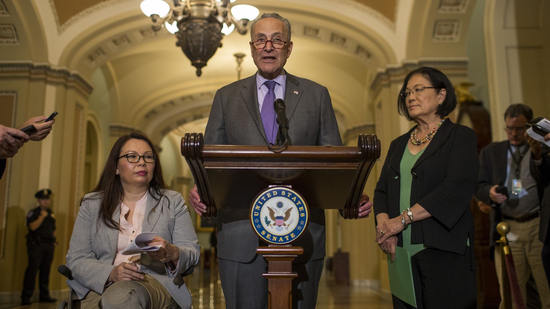 Photo of Chuck Schumer standing at a podium with Tammy Duckworth and Mazie Hirono beside him