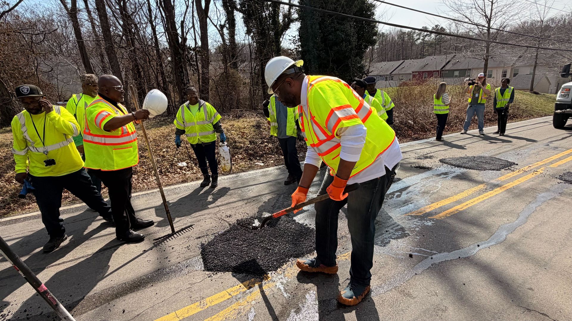Group of road workers and volunteers in neon vests repair a pothole on a sunny street. A man in a white hard hat and orange gloves shovels asphalt as others watch; a truck is parked to the right.