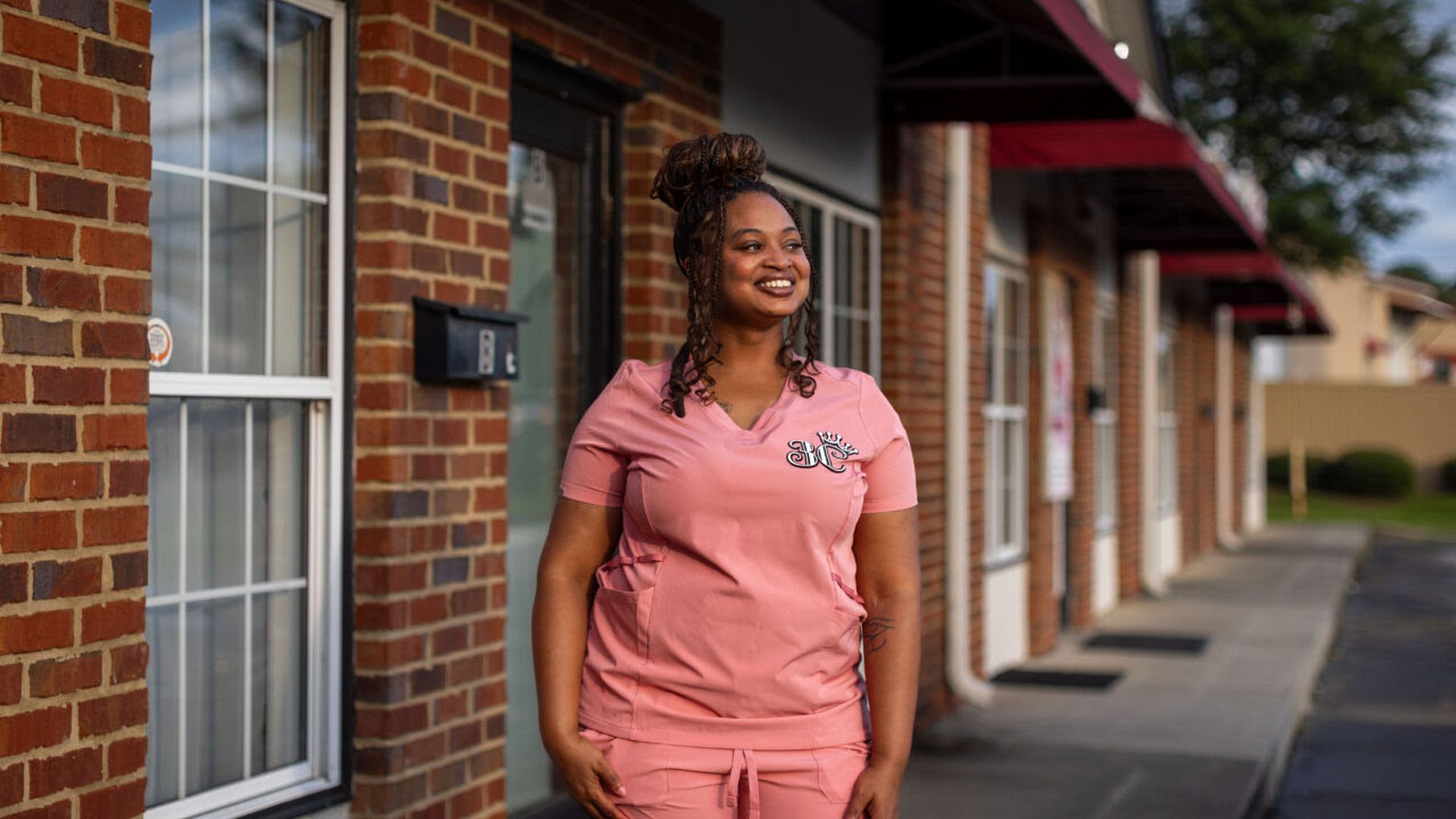 Smiling woman with braided hair wearing pink medical scrubs standing outside a brick building with windows and red awnings on a sunny day.