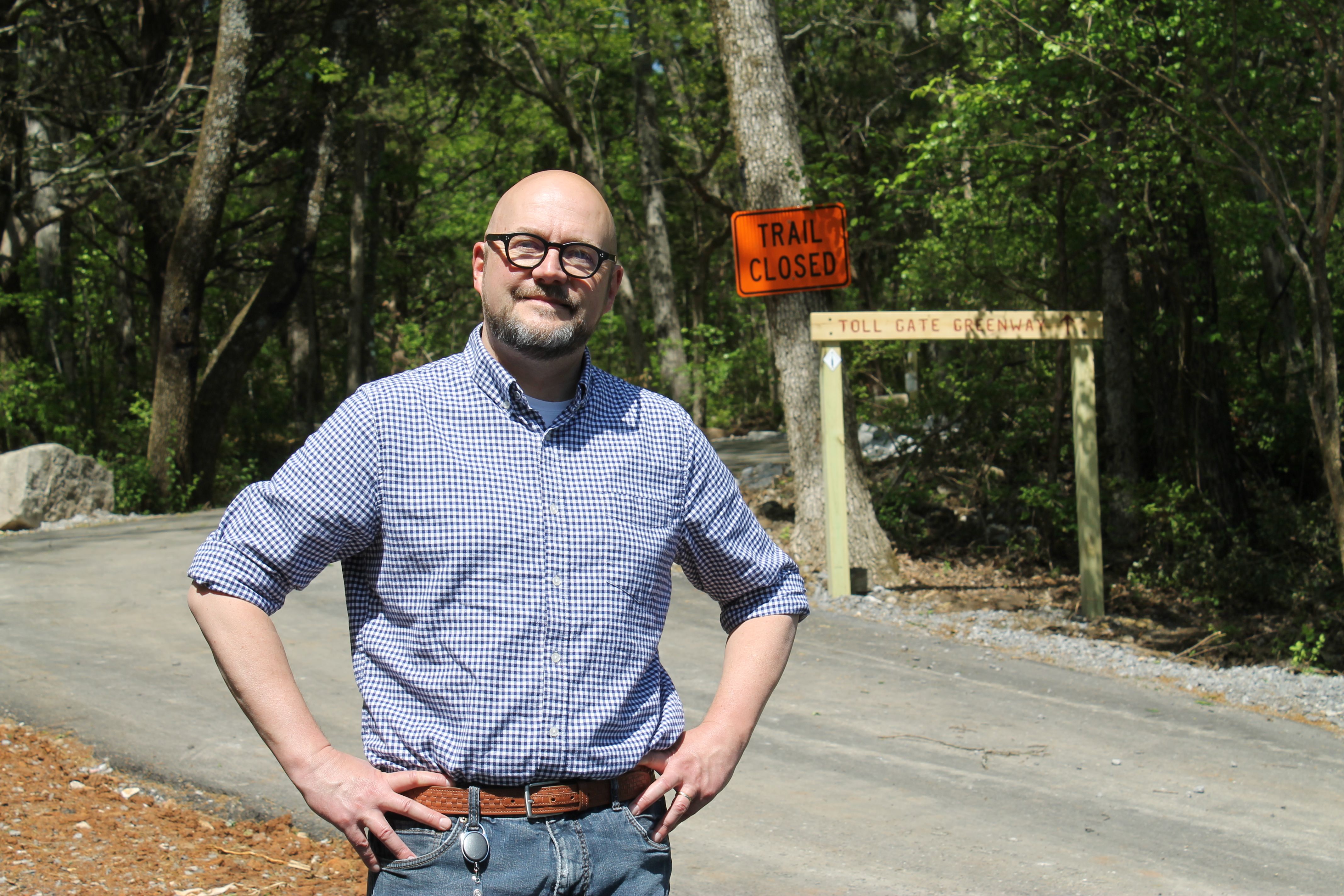 A man with glasses and a beard stands on a forest road, hands on hips, wearing a blue checkered shirt and jeans. An orange "Trail Closed" sign and a wooden gate reading "Toll Gate Greenway" behind him.
