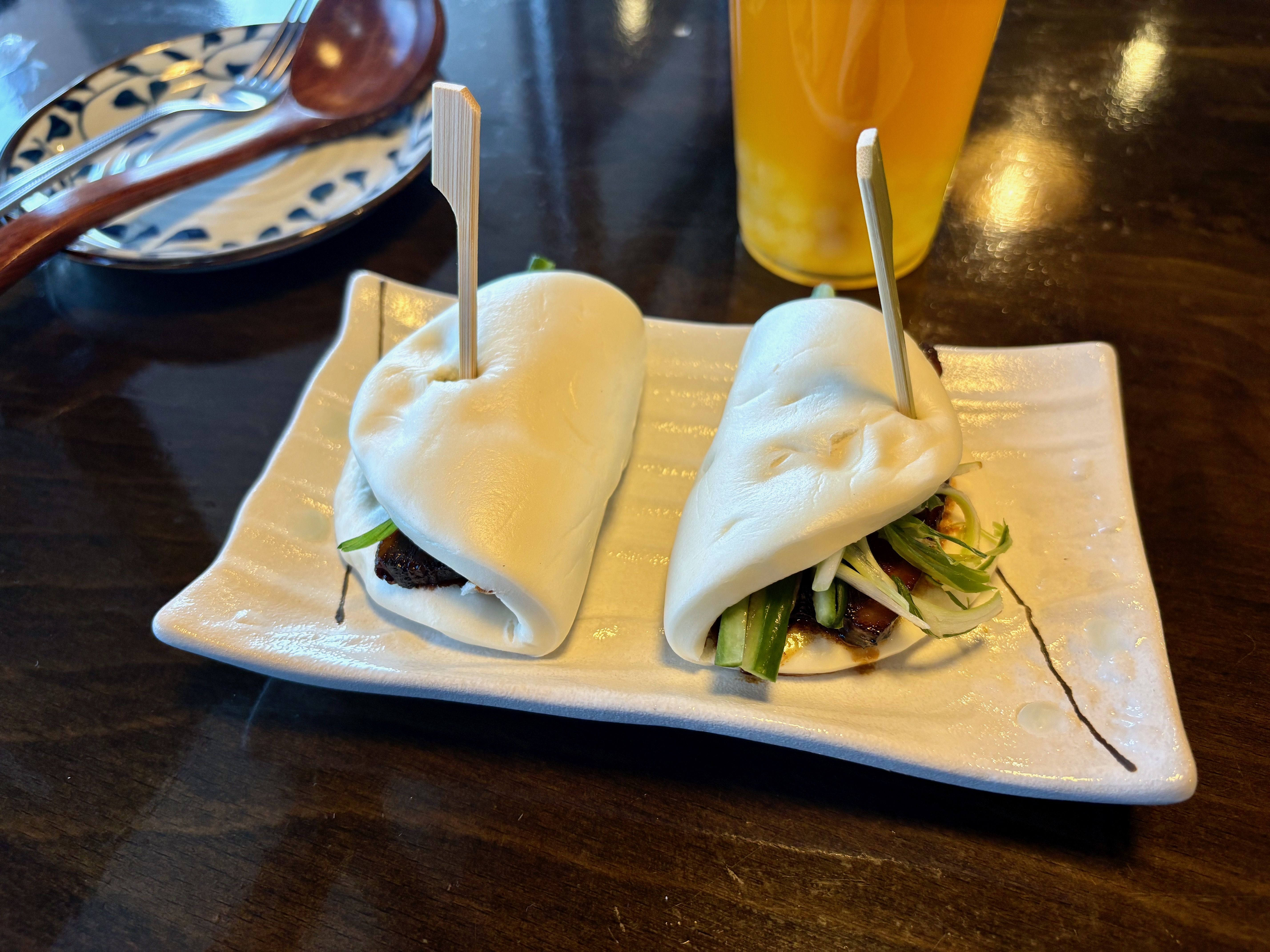 Two steamed bao buns with skewers, filled with green vegetables and meat, on a white rectangular plate on a dark wooden table, accompanied by a patterned plate and a yellow drink.