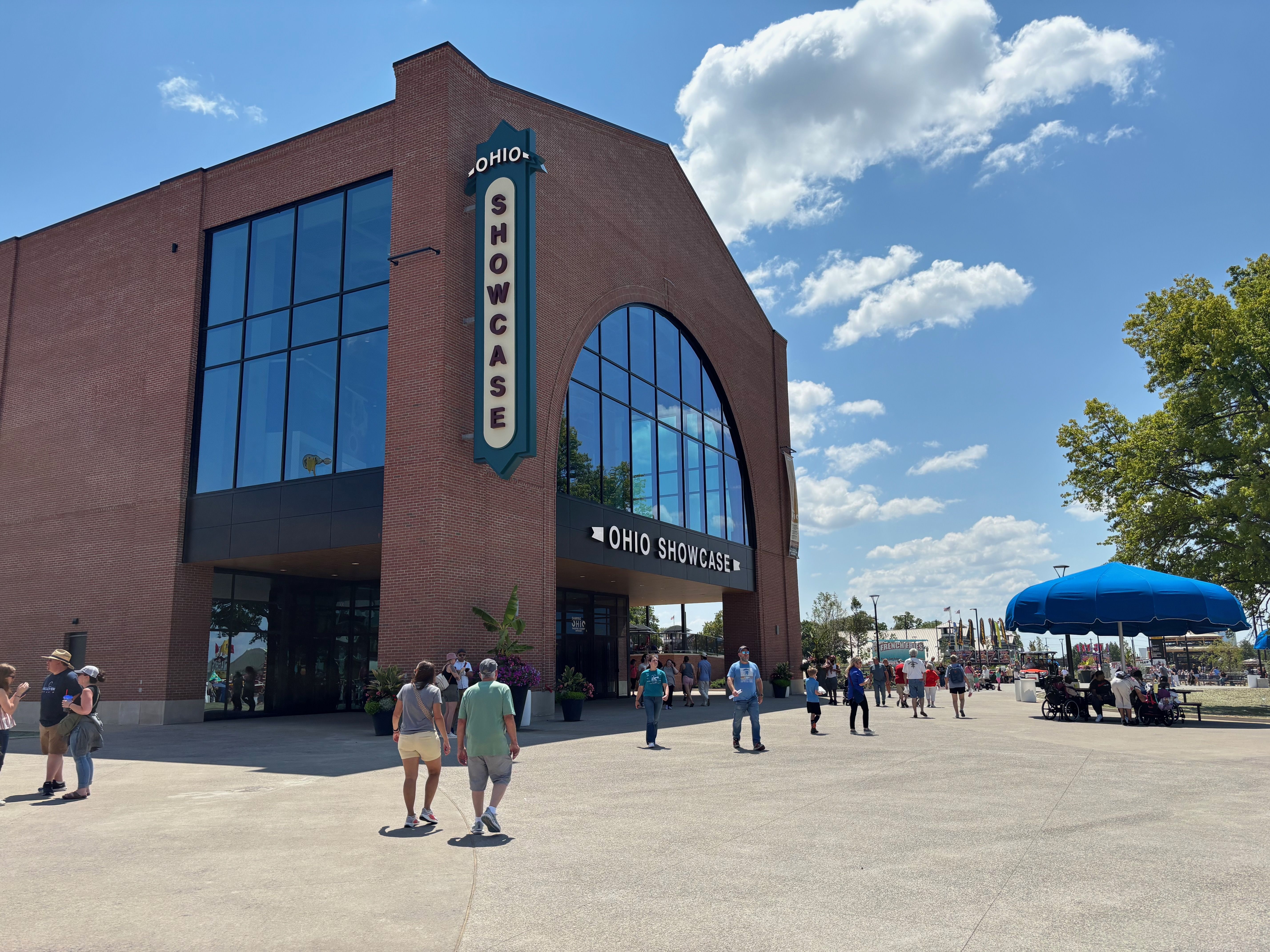 Brick building with a tall vertical sign reading "OHIO SHOWCASE" beside large glass windows. A blue sky with white clouds, and people walking outside near umbrellas and trees.