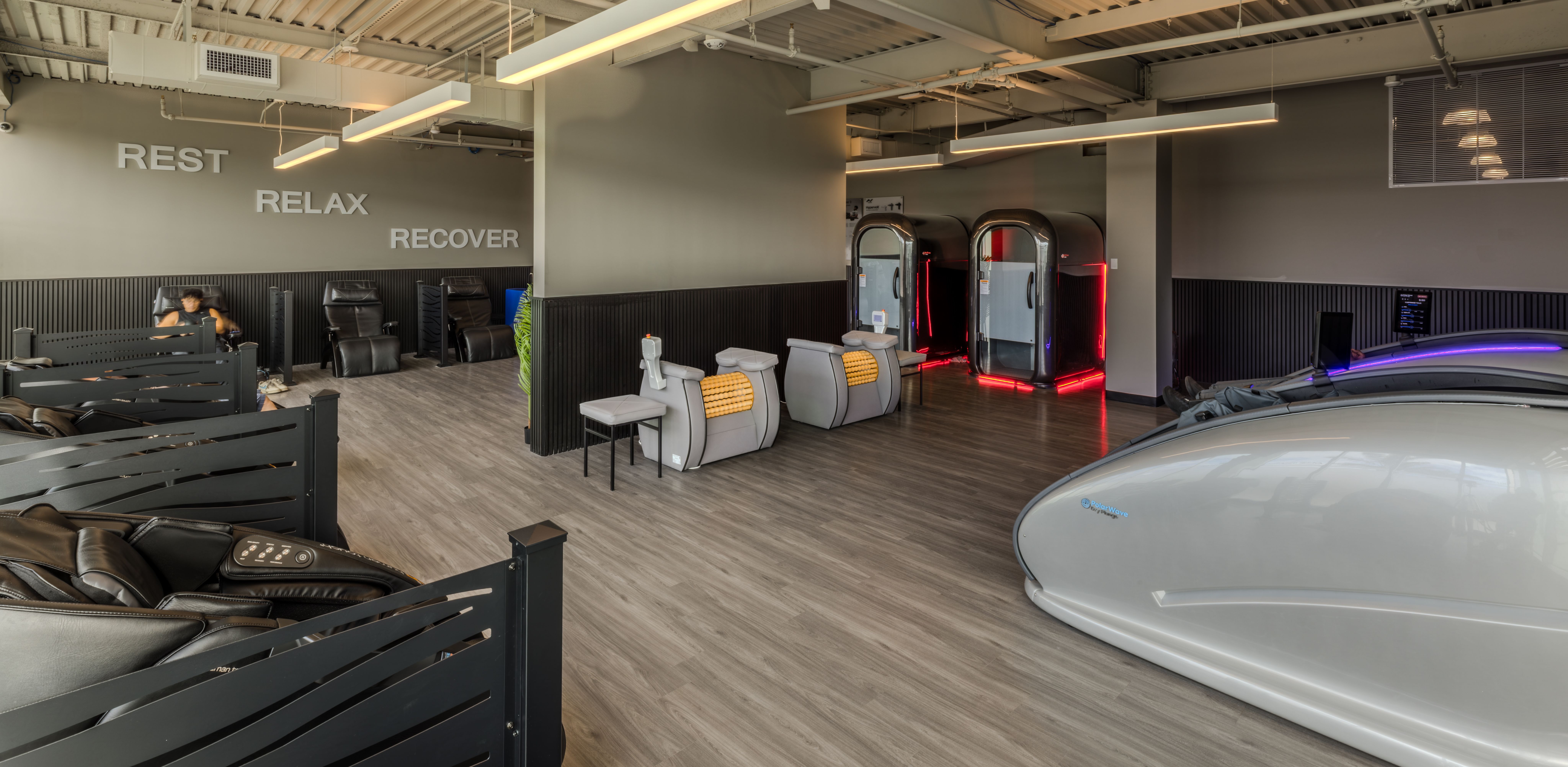 Modern wellness lounge with massage chairs along a gray wall that reads REST RELAX RECOVER, a central partition, and futuristic glass pods with red neon trim; purple lighting on the right.