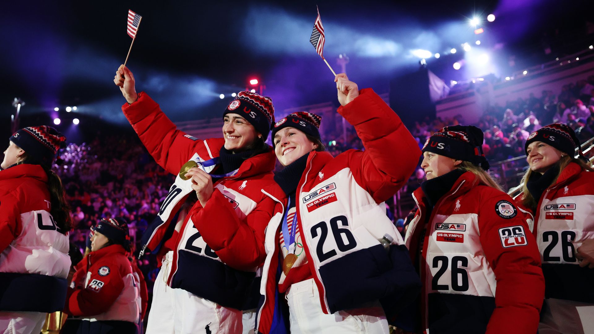 Two girls holding tiny American flags and wearing Team USA Olympic jackets in a crowd. One is also holding out her gold medal