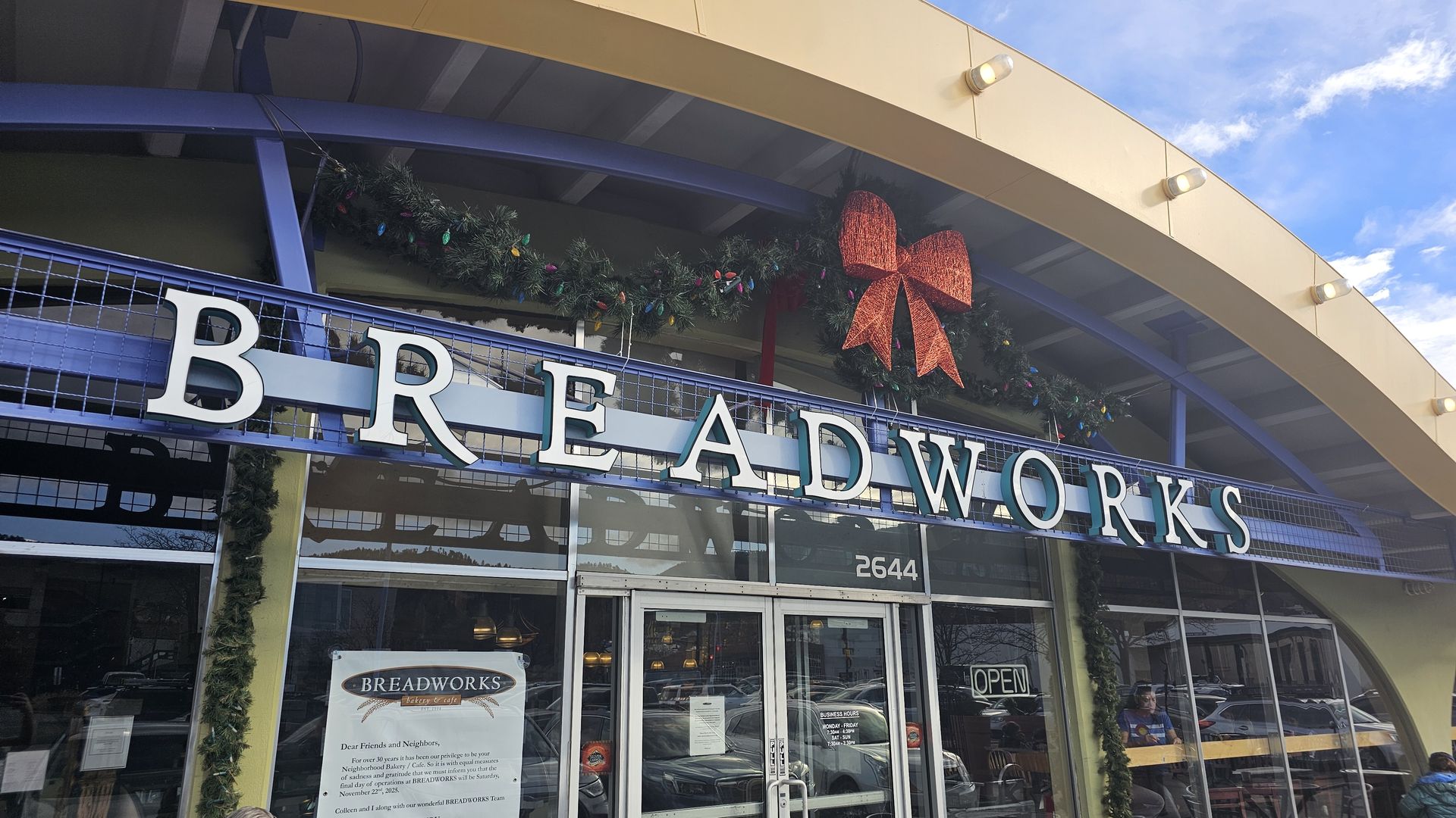 Entrance of Breadworks bakery with large white letters, holiday garland, and a big red bow above the door, under a beige and blue curved awning on a sunny day.