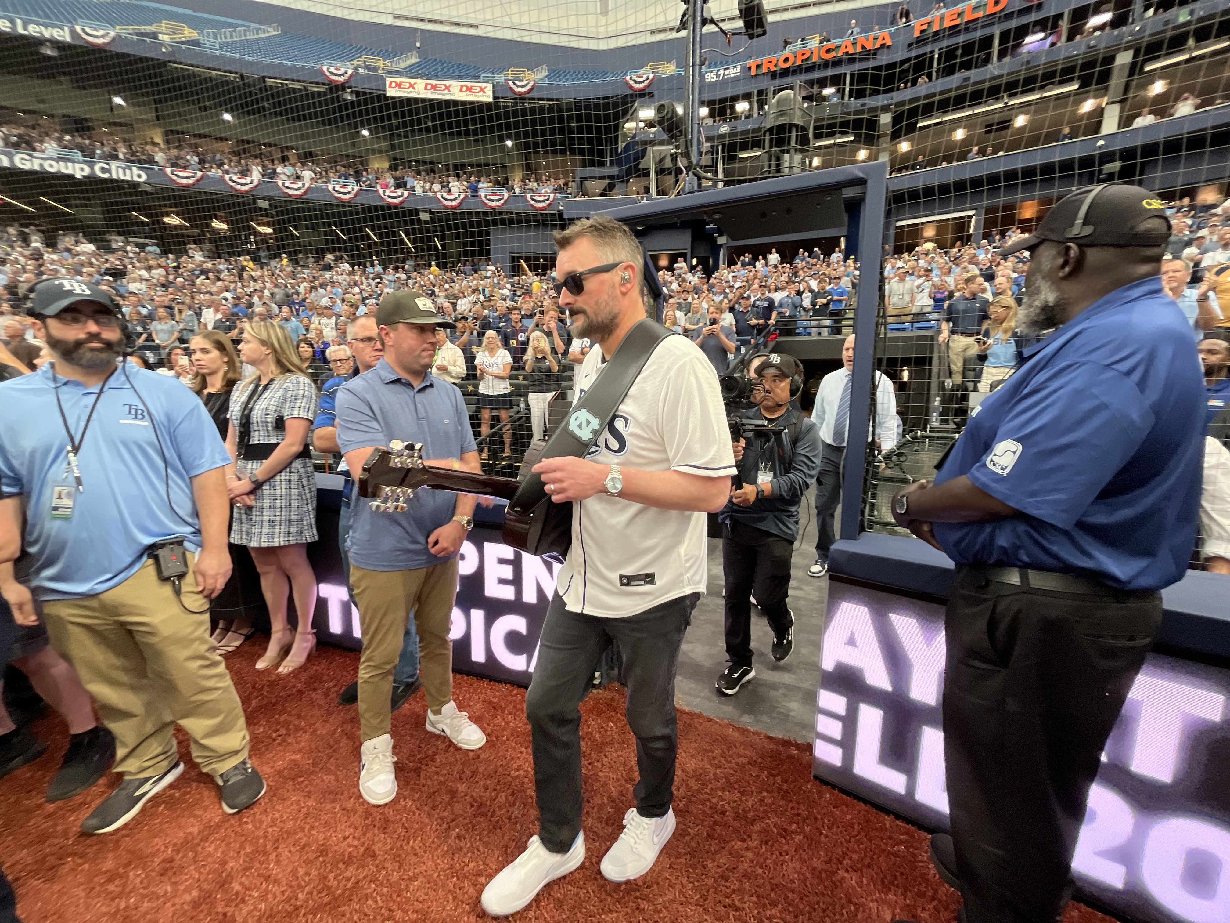 Guitarist in a white T-shirt and sunglasses walks with a guitar inside a fenced section at Tropicana Field, as staff and a large crowd watch from the stands behind a safety net.