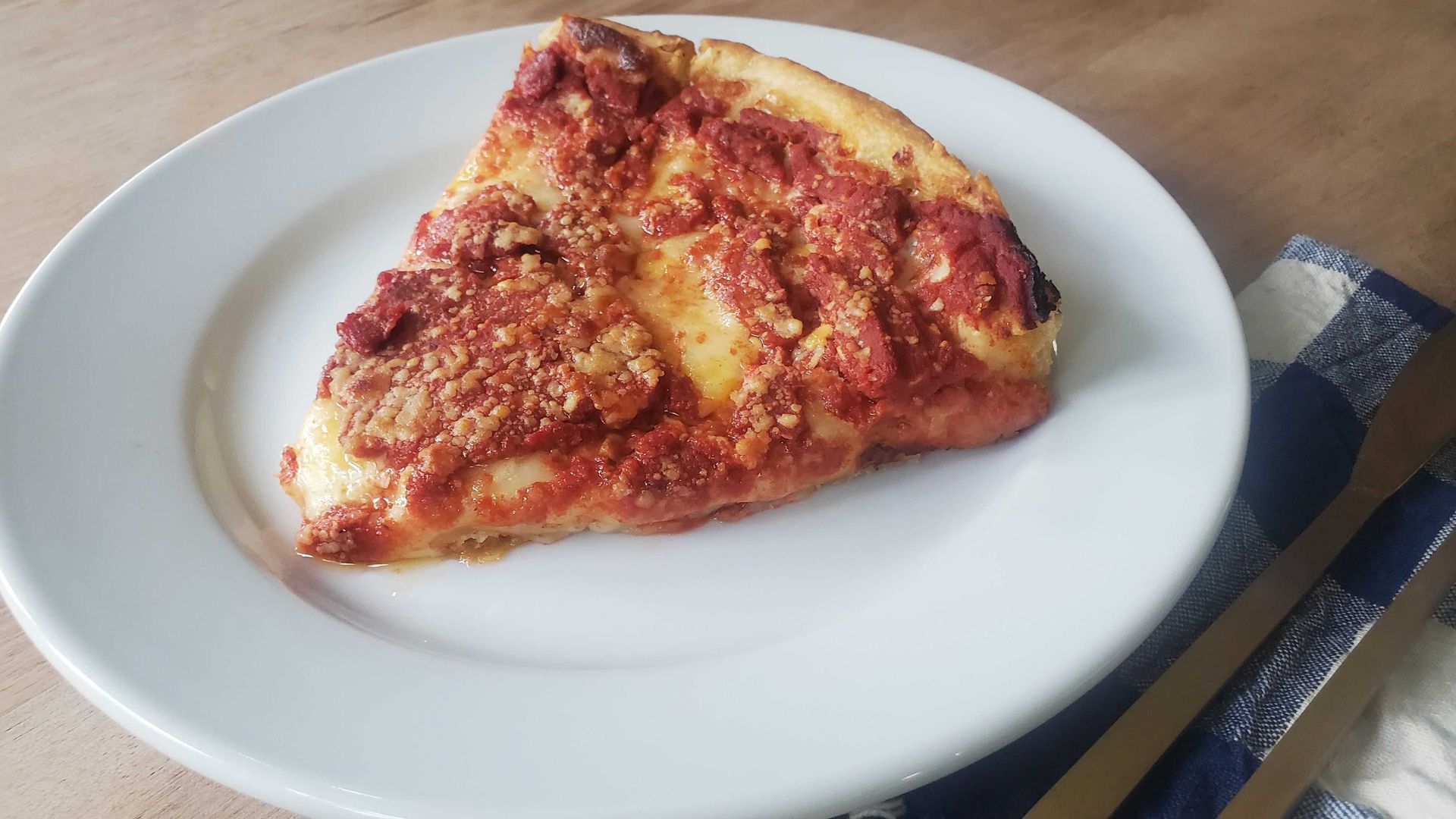 Slice of pepperoni pizza on a white plate. Tomato sauce and melted cheese with crumbled toppings, browned crust. Plate rests on a wooden table next to a blue plaid napkin and silverware.