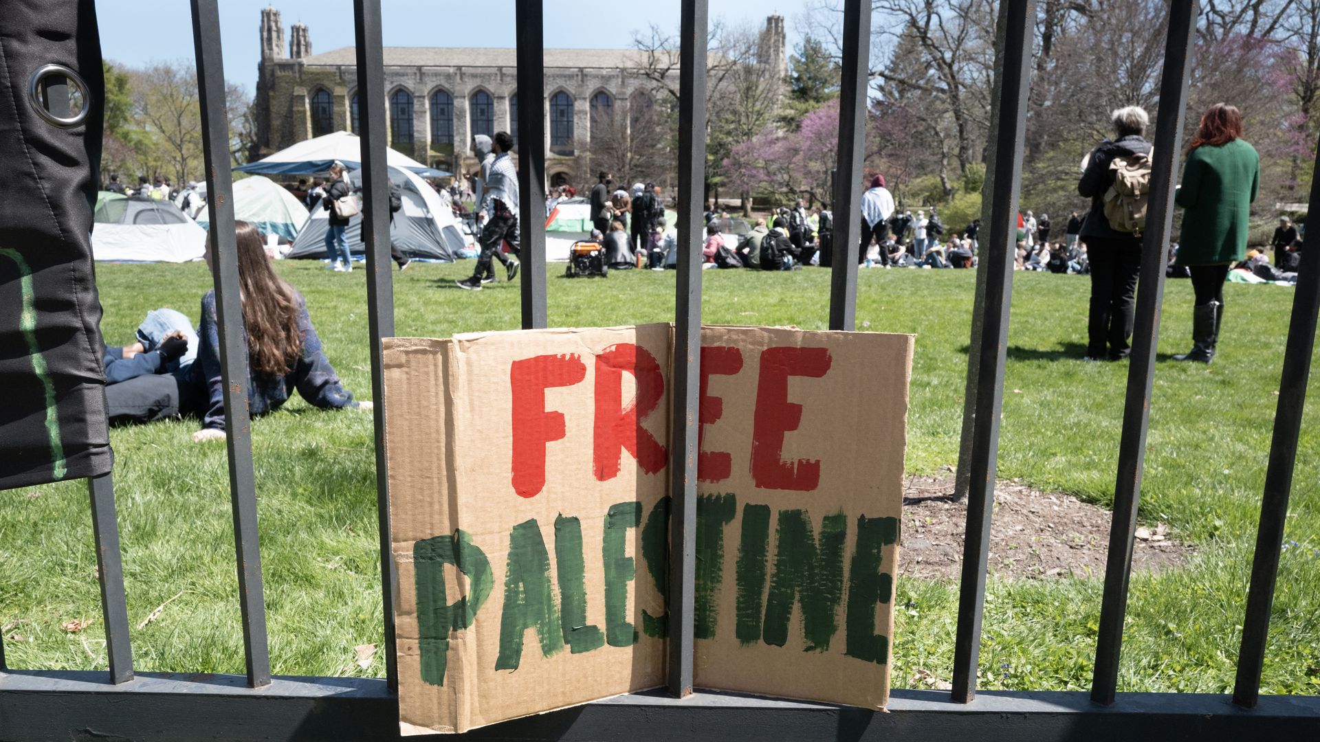 A Free Palestine protest sign sits on a metal fence with tents and protesters in front of Northwestern University in the background.