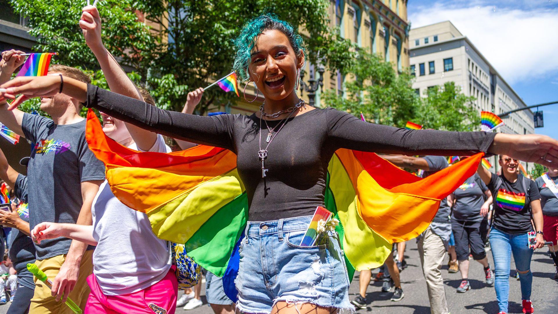 Central figure with teal hair, black top, rainbow cape, and fishnet stockings smiles as a parade winds through a sunny city, rainbow flags waving.