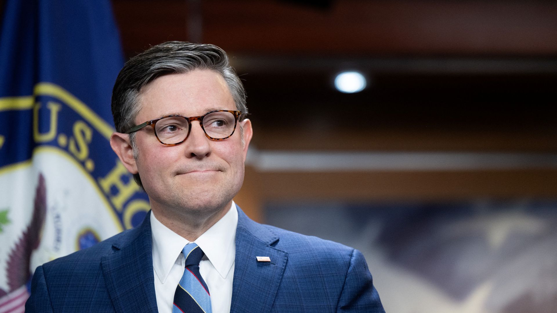Mike Johnson — wearing a blue suit, a white collared shirt, a striped blue tie and brown glasses — looks on during a press conference.