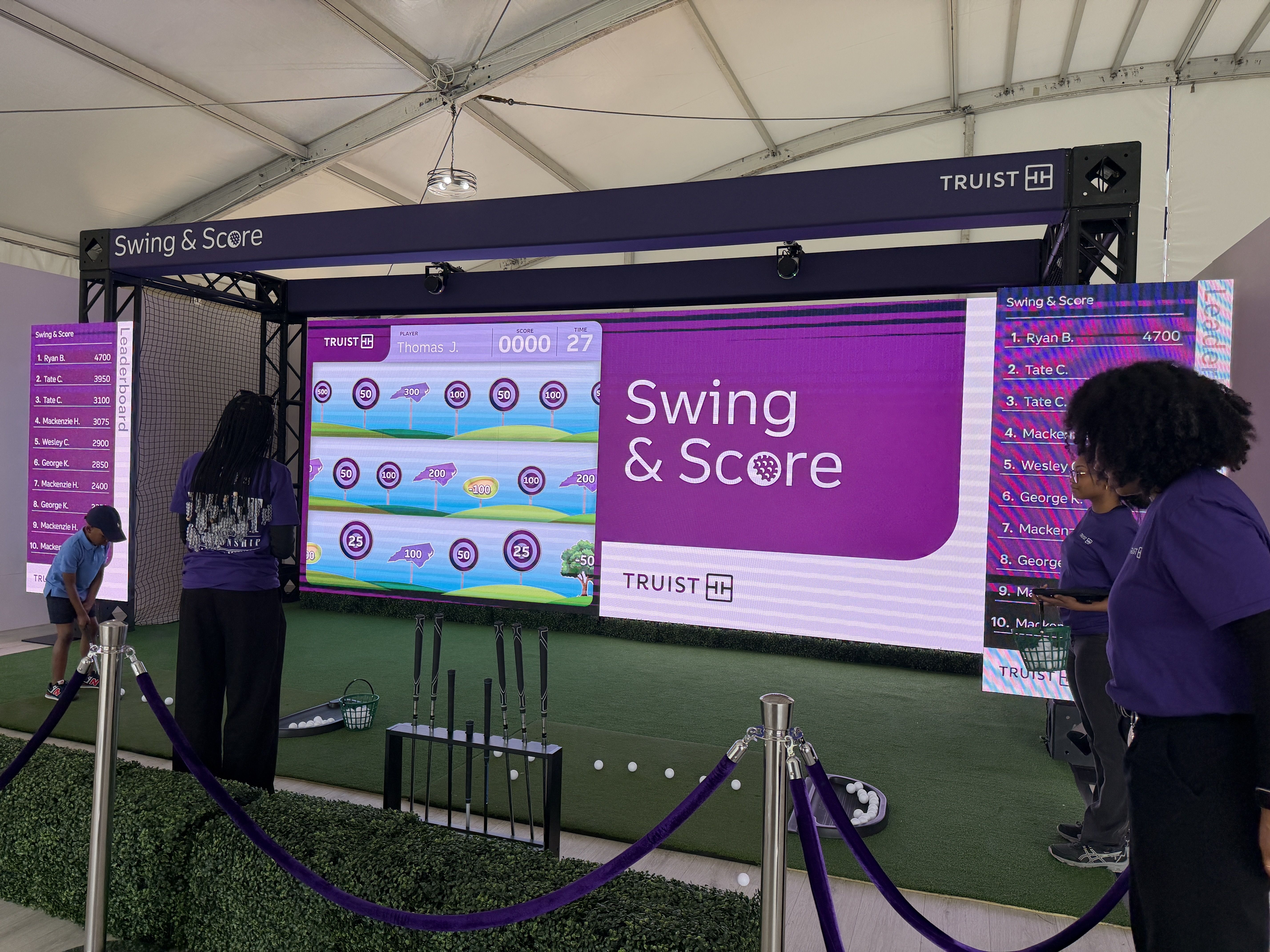 Purple-themed Swing & Score booth inside a white tent, with a large screen golf game, side leaderboards, a metal frame, netting, and people in purple shirts nearby.