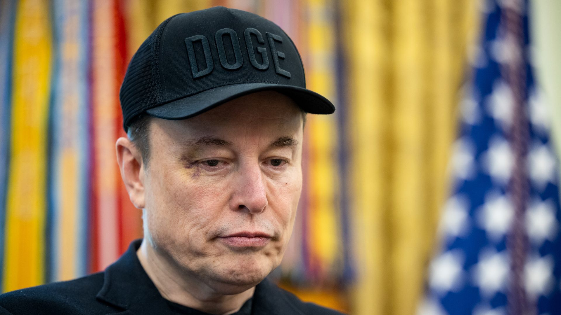 Elon Musk wearing a black DOGE hat and standing in front of a gold curtain and an American flag.