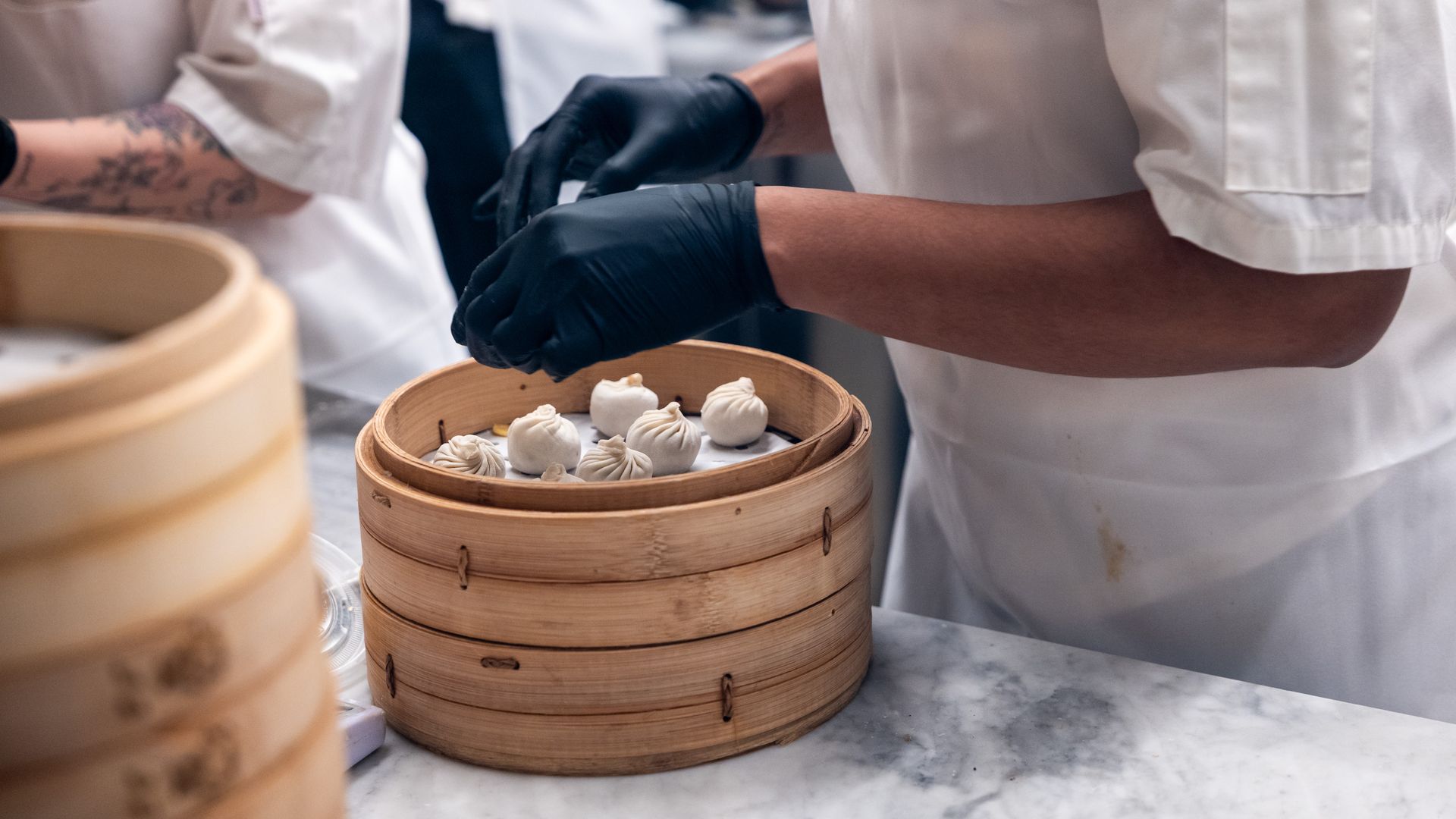 Two chefs in white uniforms and black gloves work together to assemble small dumplings in a wooden bamboo steamer, placing delicate pastries on a marble counter.