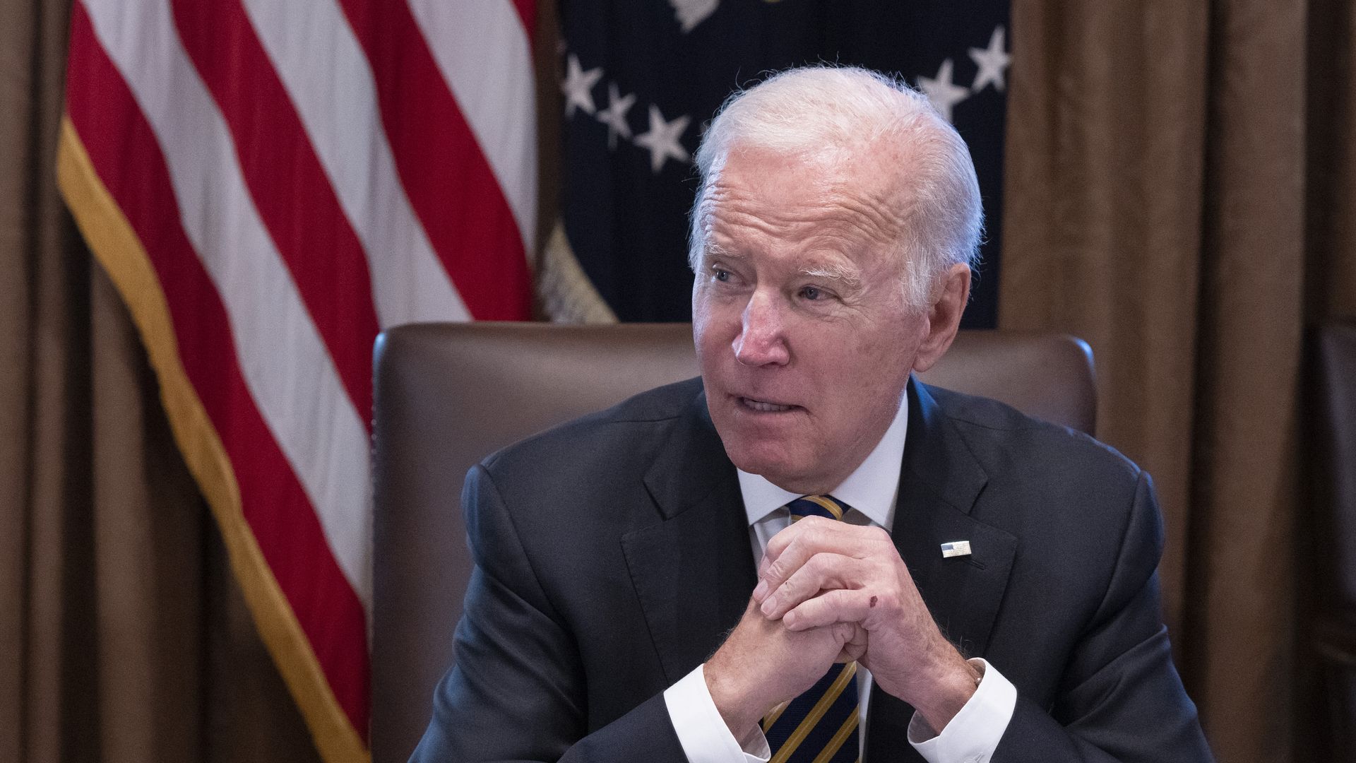 Photo of Joe Biden sitting at a table with his hands clasped in front of him as he looks to his right