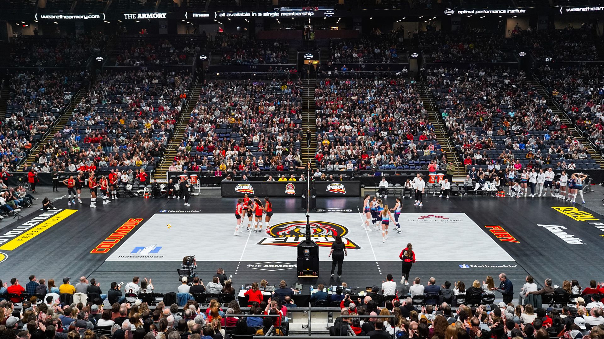 Indoor volleyball match with large crowd, players in red and blue uniforms on court, and logos including Columbus Fury and Nationwide on the floor.