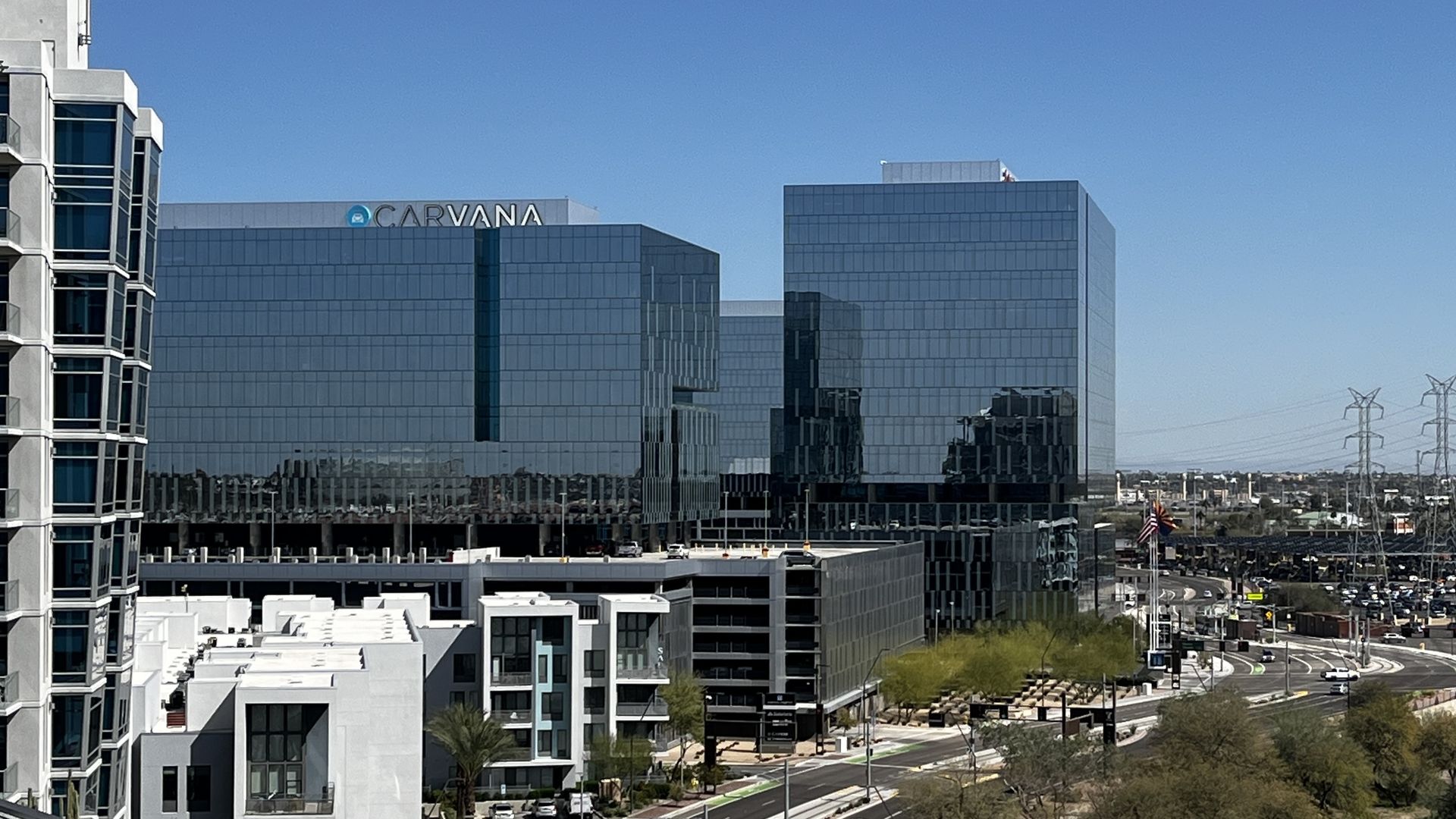 Cityscape with modern glass office buildings, including one with "CARVANA" signage, white residential buildings, a curved road, green trees, and power lines under a clear blue sky.