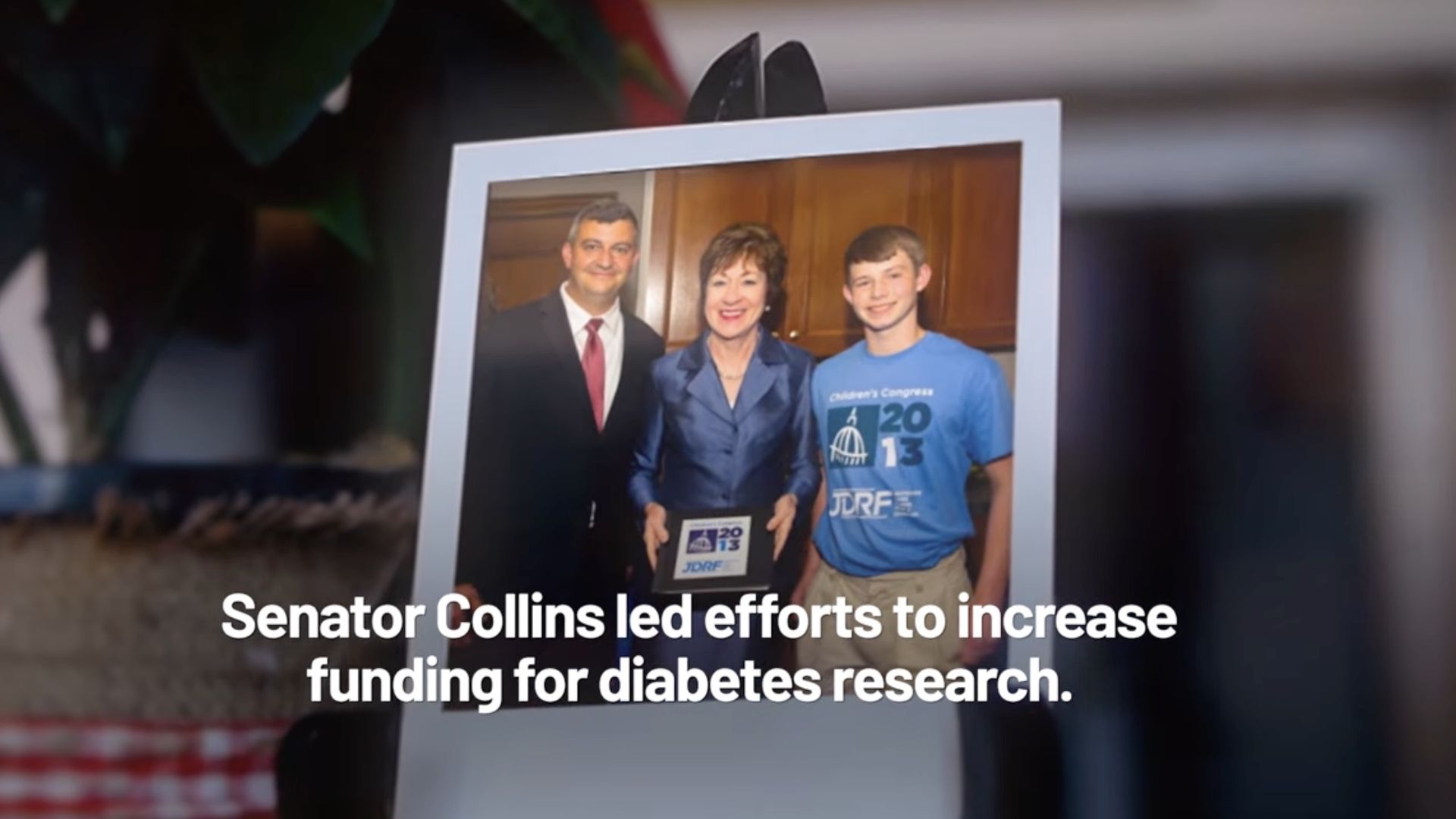 Framed photo of three people indoors: a man in a dark suit with a red tie, a smiling woman in a blue suit, and a young man in a blue T-shirt with '2013' and JDRF logos.