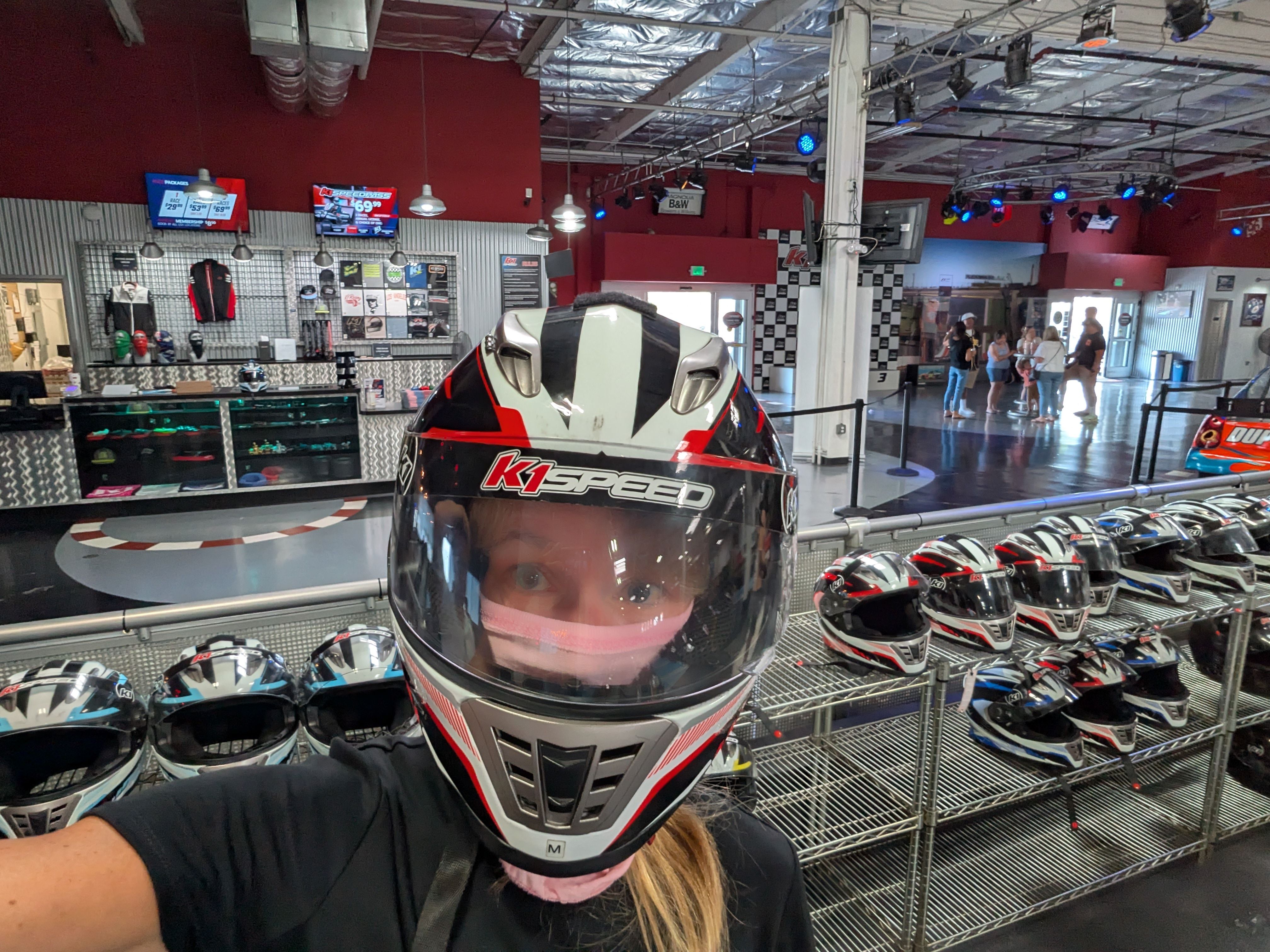 Selfie inside a K1 Speed go-kart shop: person in a white, red, and black full-face helmet foreground; shelves with helmets and racing gear, blue stage lights, and a track area in the background.