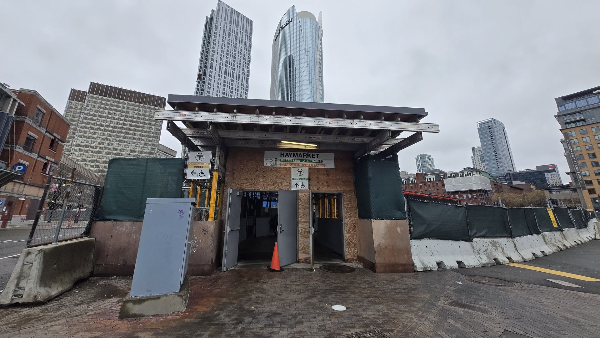 Urban construction site with a shelter at a tunnel entrance labeled Haymarket, plywood walls, orange cone, brick paving, green fencing, and tall modern skyscrapers under a cloudy sky.