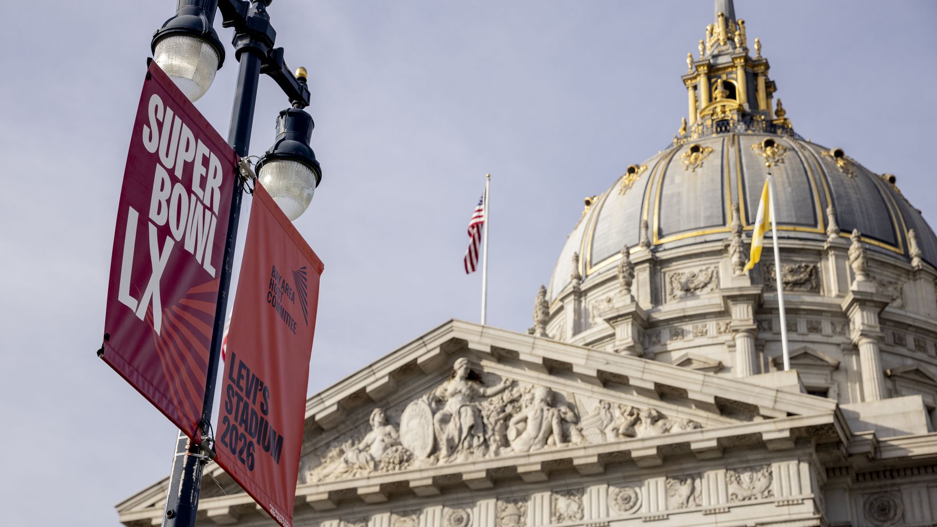 Red banners on a street lamp advertise Super Bowl LX and Levi's Stadium 2026, with an ornate domed building featuring a flagpole and American flag in the background under a clear sky.